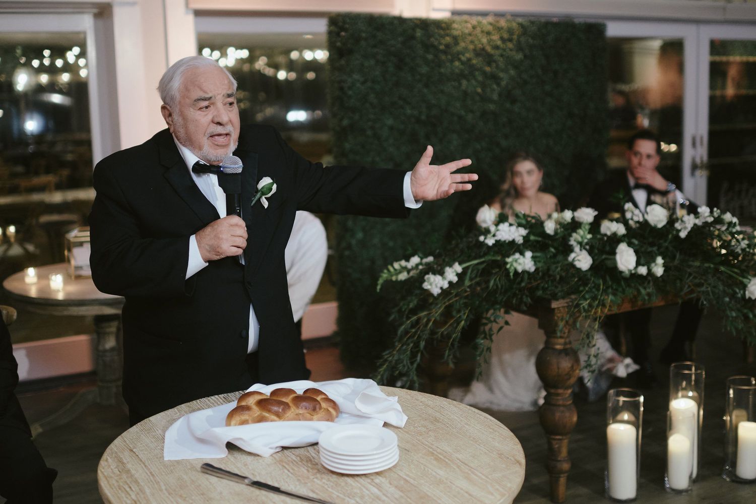 A speaker gives a toast at an elegant wedding reception with white floral decor and candlelight.