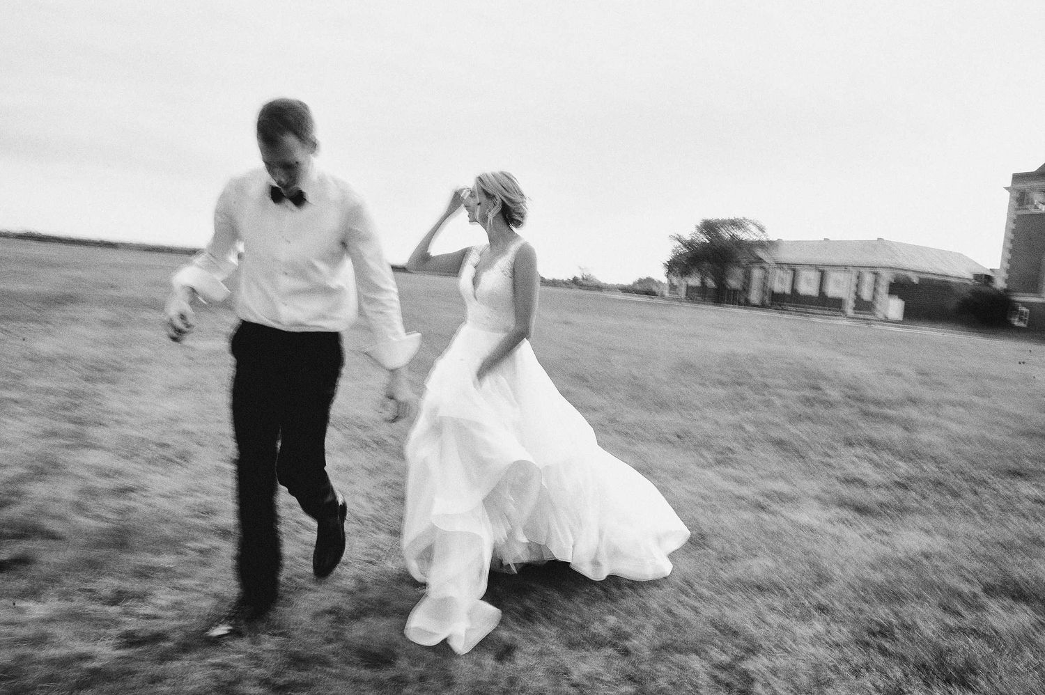 A candid black and white photo of newlyweds running together across an open field.