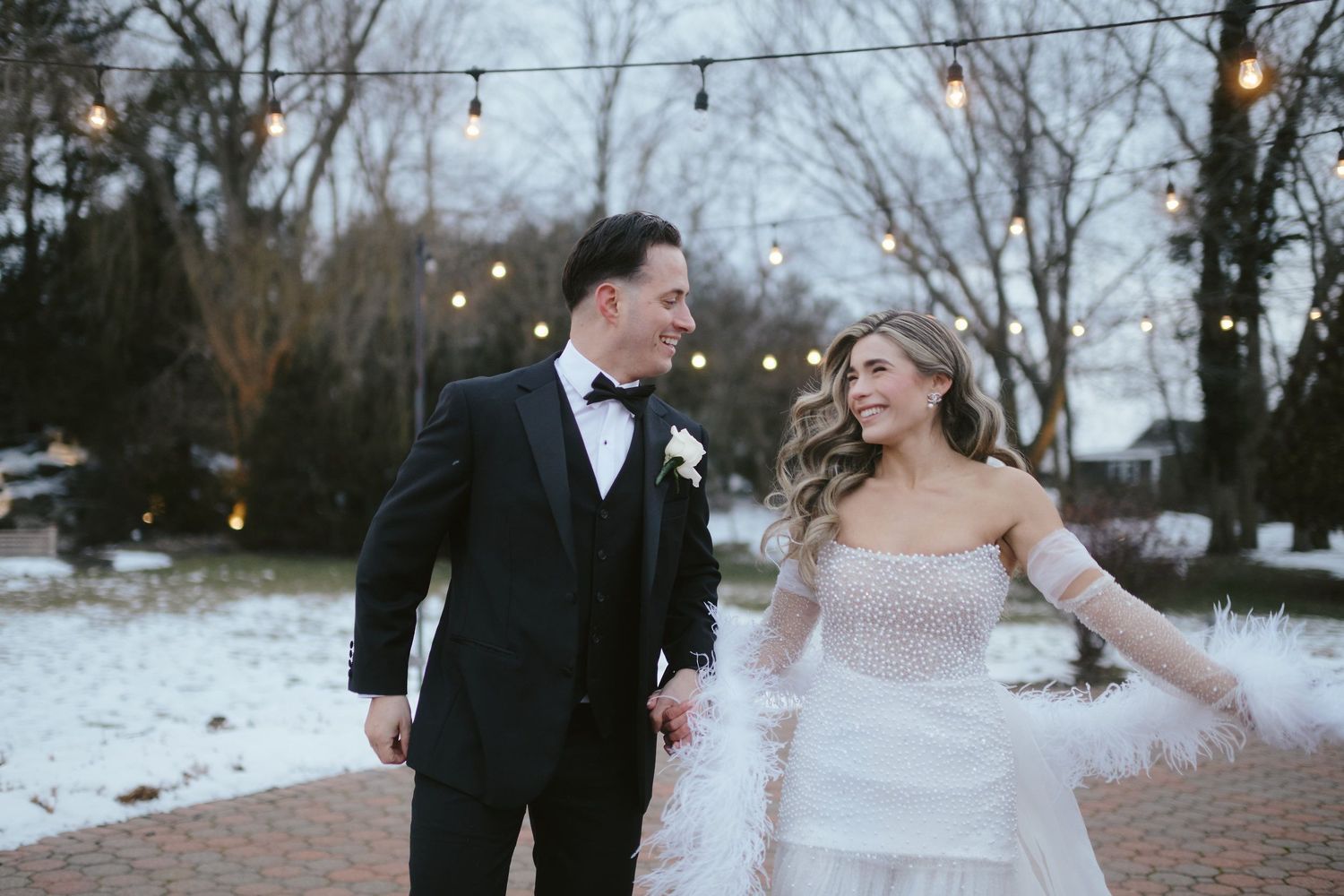 A joyful couple celebrates their winter wedding day on a snow-covered brick path at dusk.