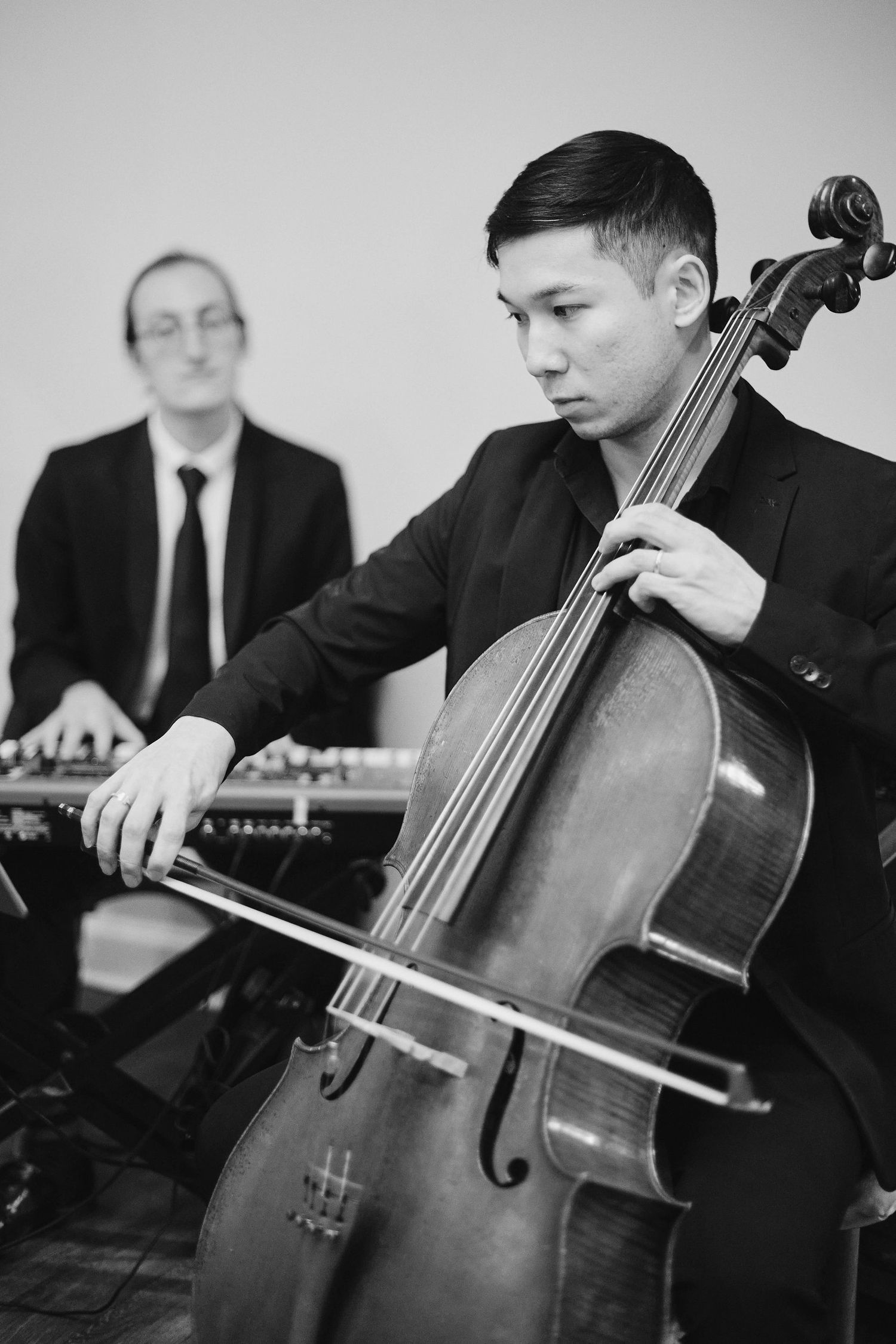 Black and white photo of musicians performing together, with a cellist in the foreground and pianist in the background.