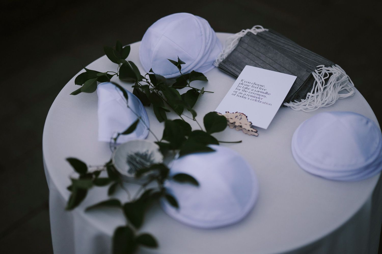 White kippahs and greenery arranged on a table with a prayer card for a Jewish wedding ceremony.