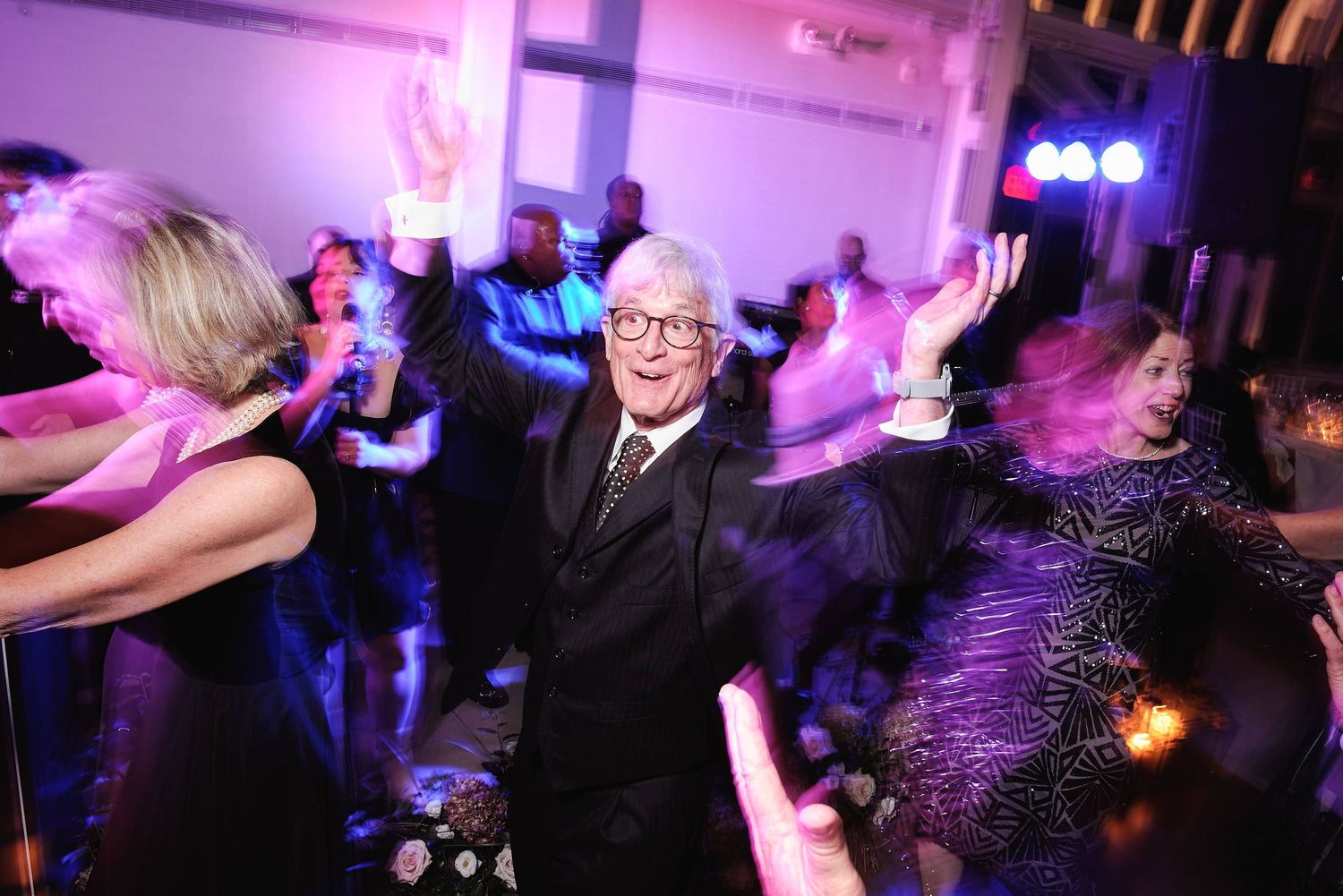 Energetic partygoers dancing and celebrating on a dance floor illuminated by purple and blue lighting at a wedding reception.