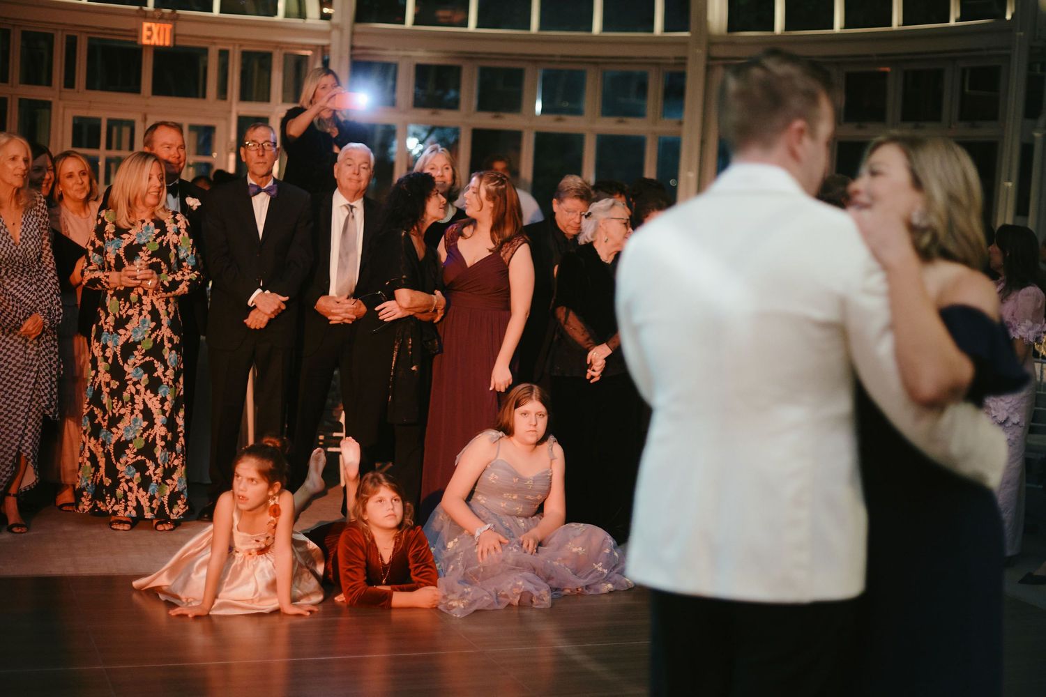 Wedding guests watch couples dancing while bridesmaids in pink dresses sit on the dance floor at an evening reception.