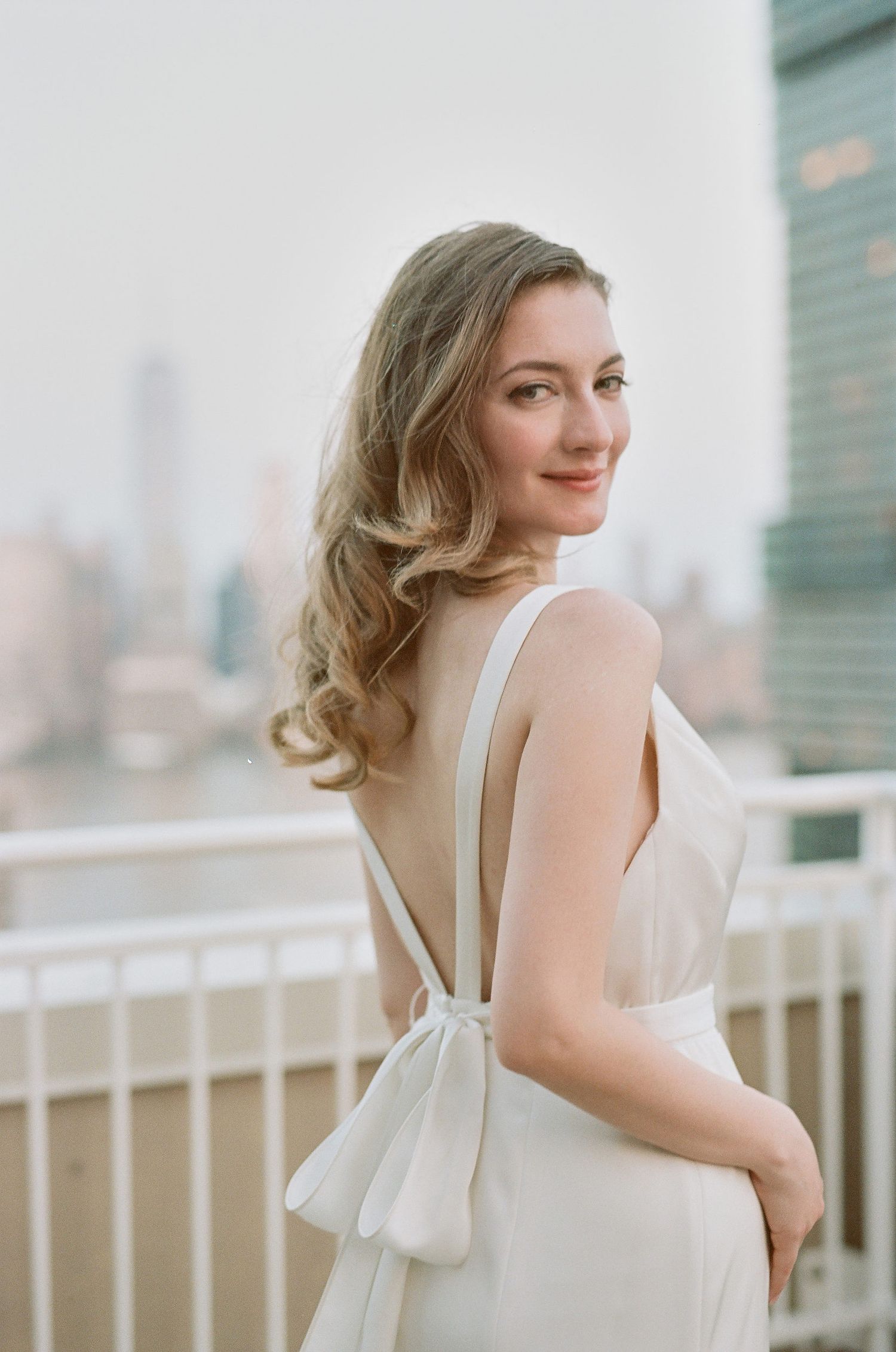 A woman in an elegant white dress poses on a rooftop terrace with city skyscrapers in the background.