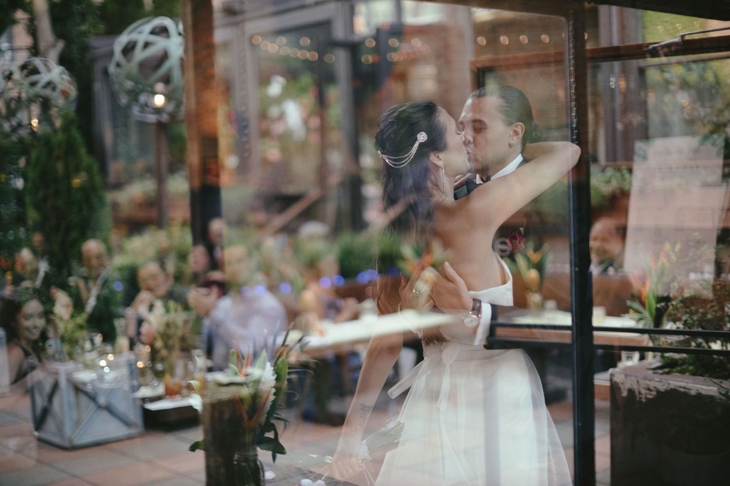 A couple share an intimate moment during their wedding reception in a glass-walled venue surrounded by greenery.