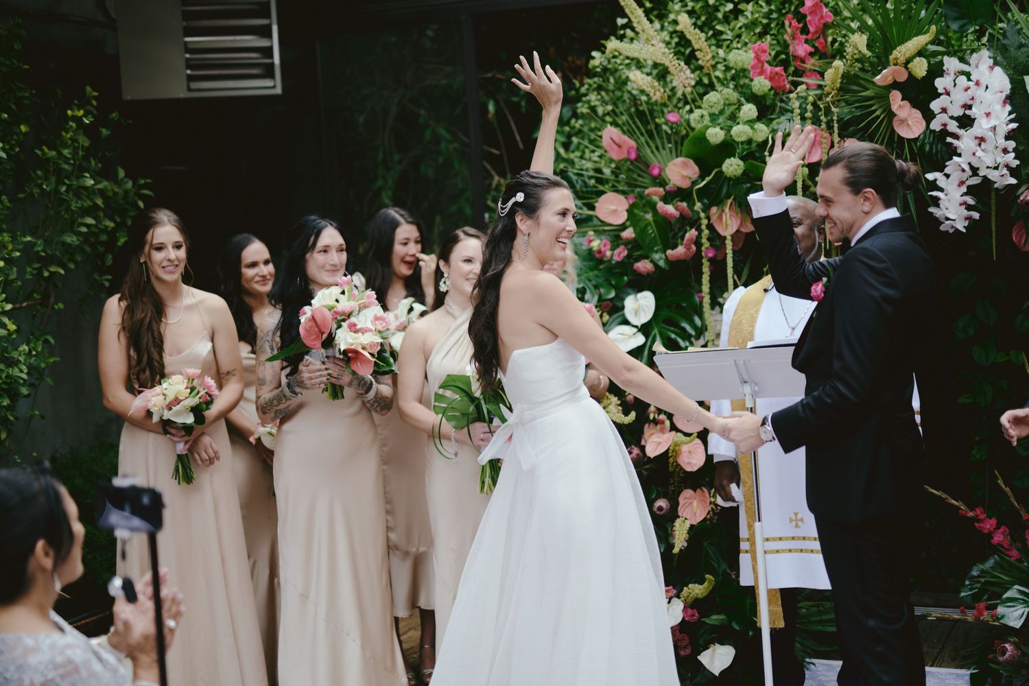 An outdoor wedding ceremony takes place in a garden setting with blush pink florals and natural greenery.