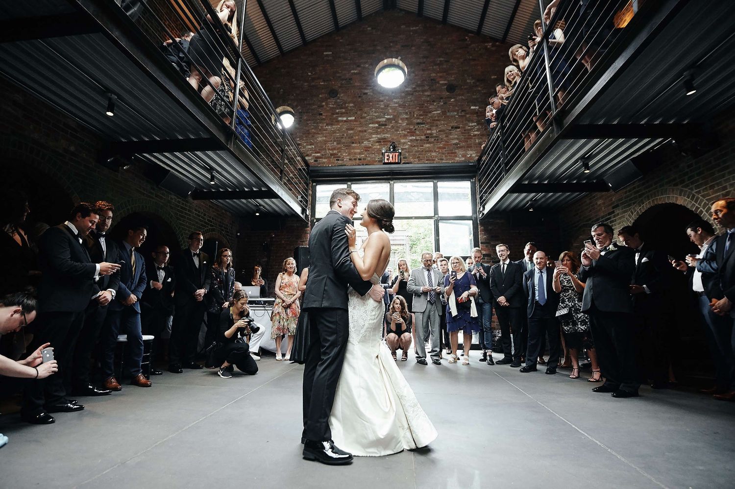 A couple shares their first dance surrounded by wedding guests in a rustic industrial venue with brick walls and high ceilings.