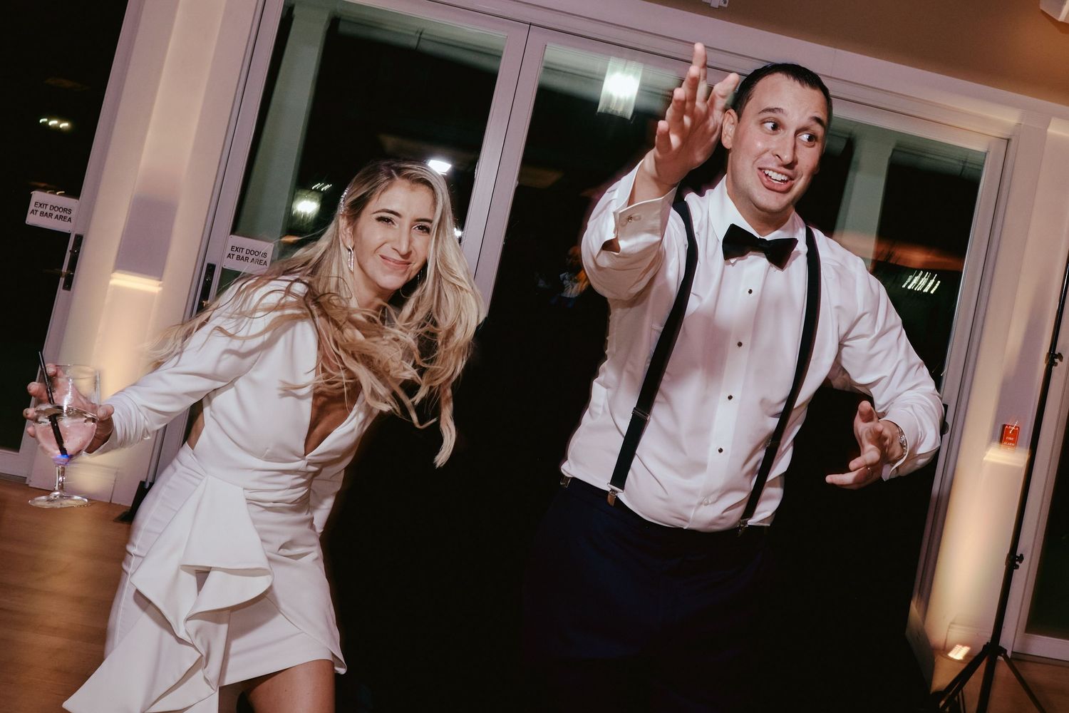A bride and groom dance playfully and energetically at their wedding reception, both wearing white and suspenders.