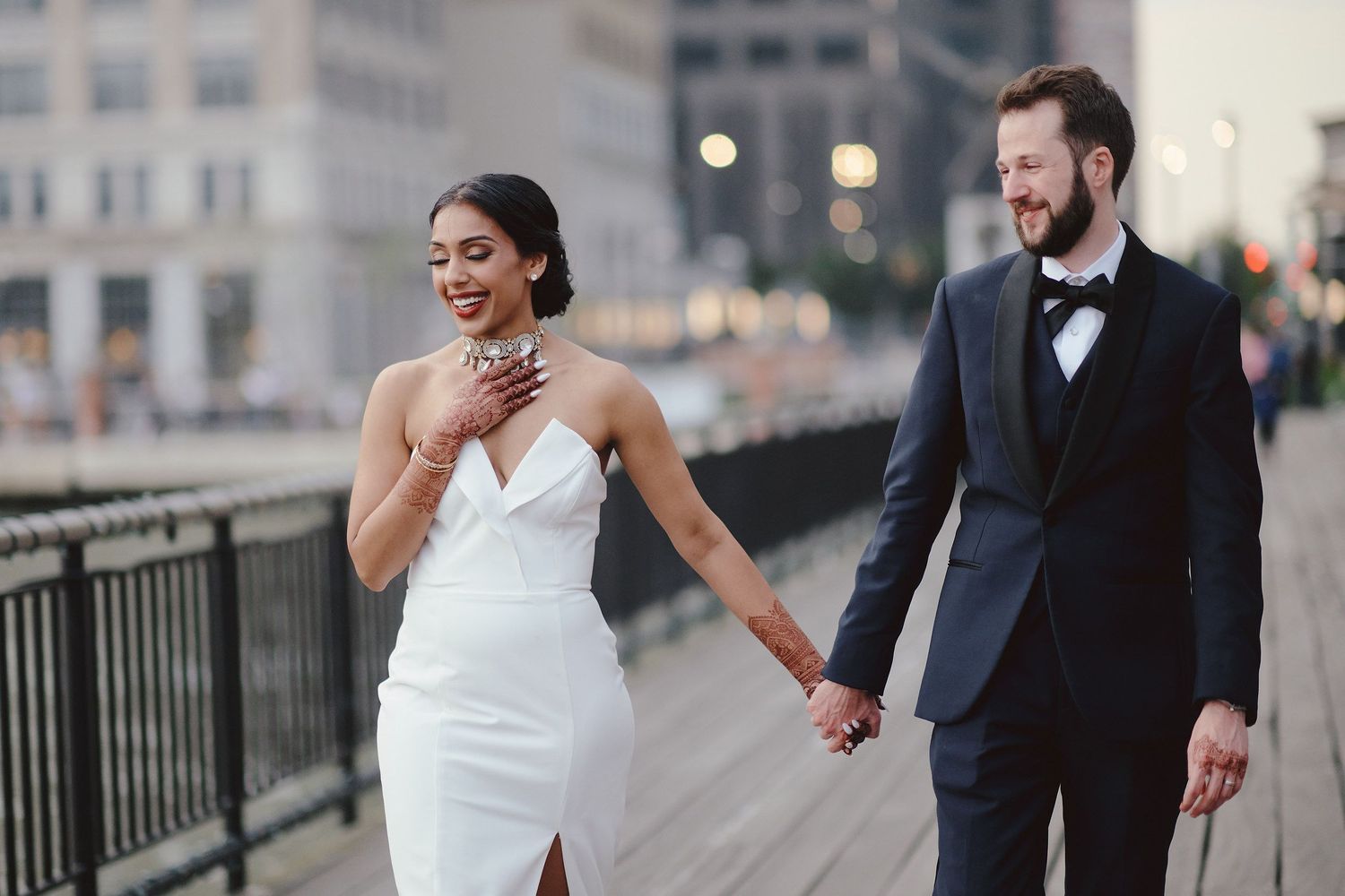 A stylish wedding couple whimsically walks along an urban railing in classic formal attire at dusk.