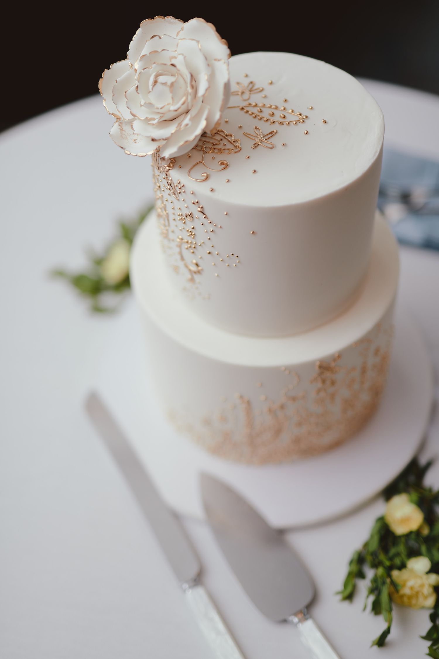 Elegant two-tier white wedding cake decorated with a white sugar rose and delicate gold accents on a white plate.