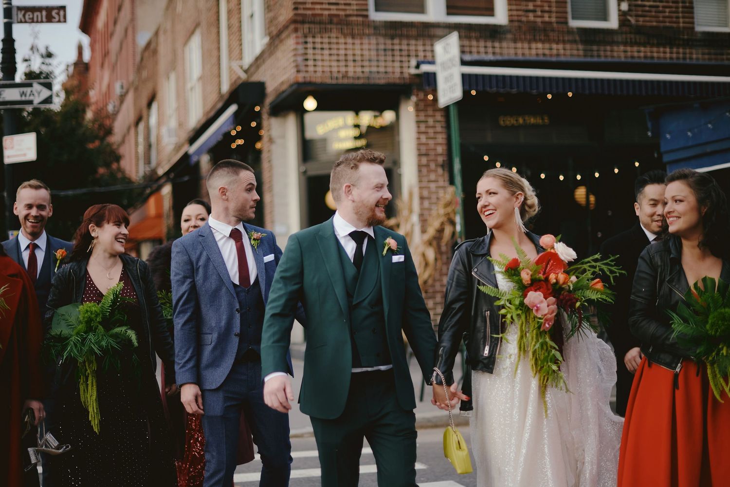 Wedding party walking along city sidewalk with bridesmaids in red and green dresses and long fern bouquets.