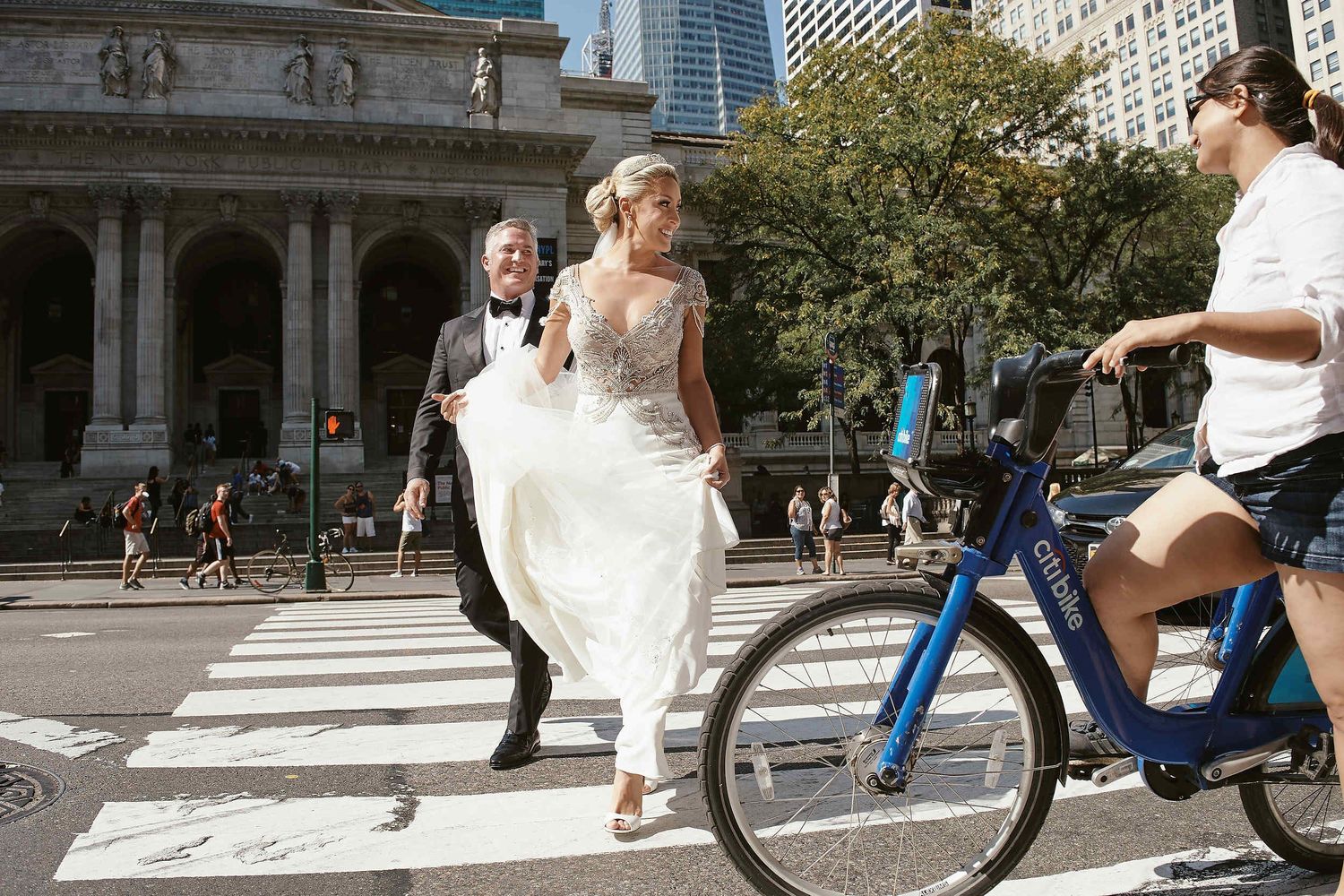 Person in wedding dress crossing city street while cyclist passes by near historic building.