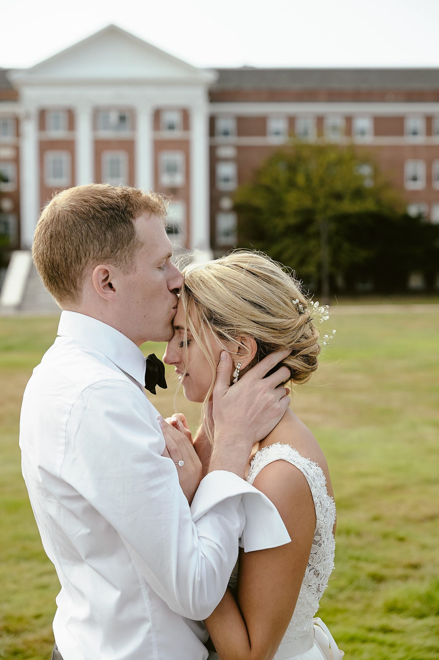 A tender wedding moment captured outside a stately brick building on a sunny afternoon.