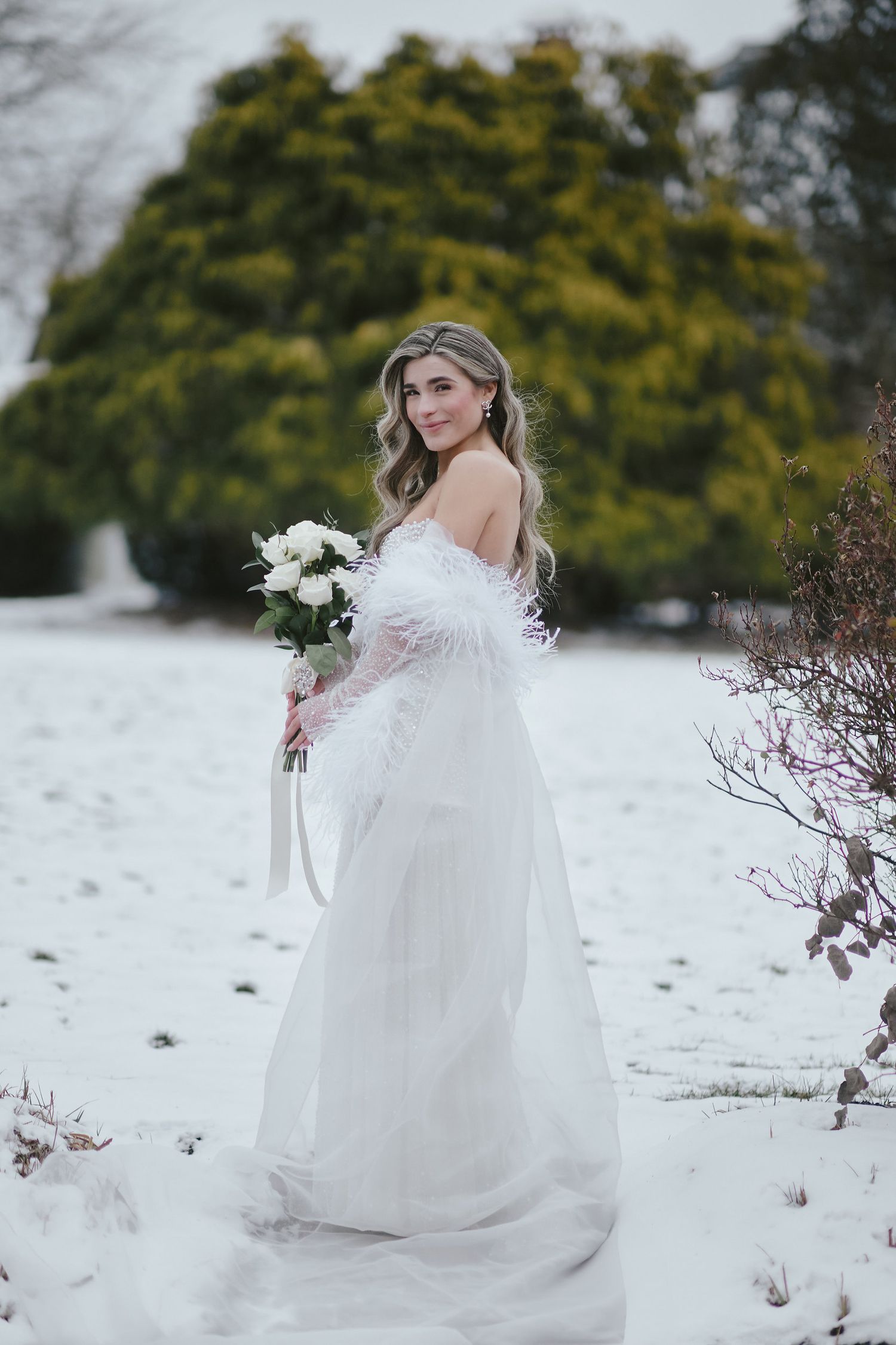 A bride in a flowing white off-shoulder gown holds a bouquet while standing in fresh snow with evergreens behind her.