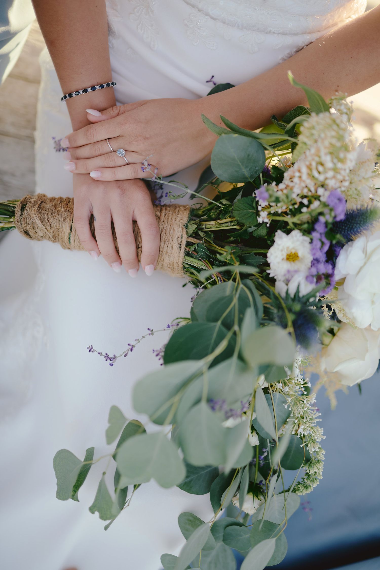 Close-up of hands holding elegant bridal bouquet with white flowers, eucalyptus leaves and purple accents.