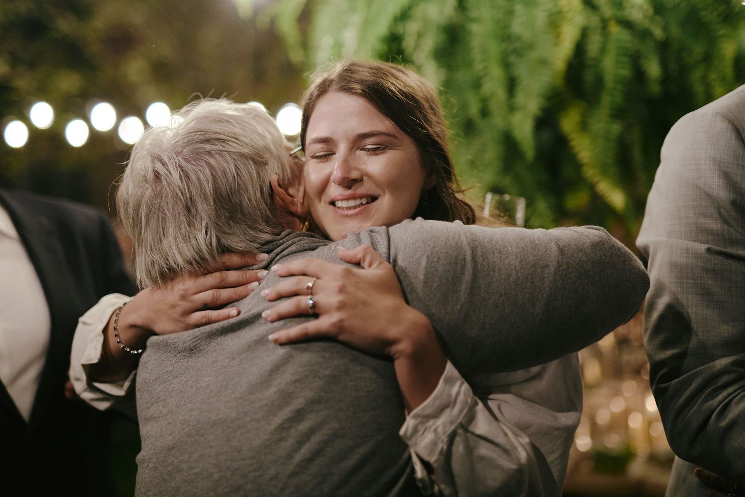 Two people share an emotional embrace at an outdoor evening gathering with greenery in the background.