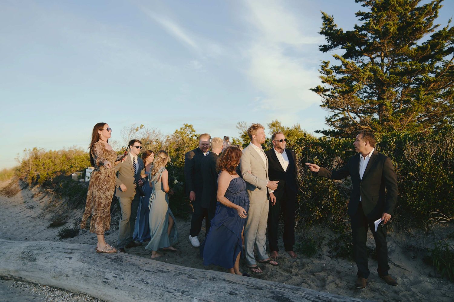 A group of formally dressed guests gather outdoors on a sandy path near evergreen trees at sunset.