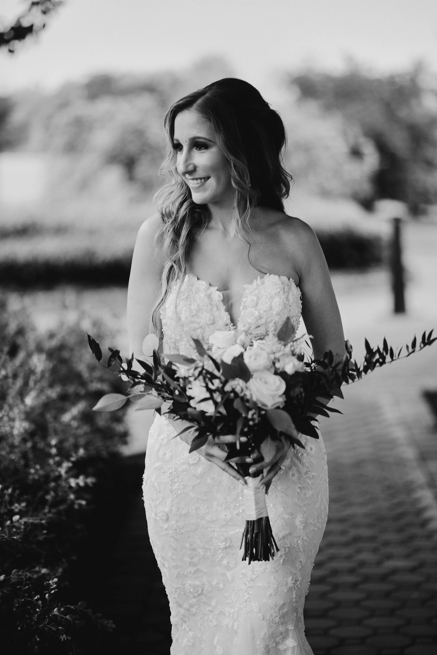 Black and white wedding photo showing bride in lace strapless gown holding eucalyptus bouquet outdoors.