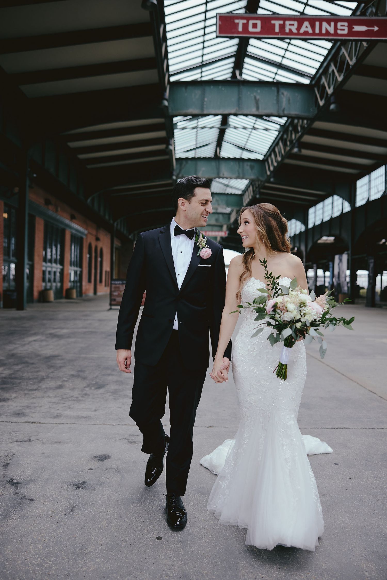 A bride and groom walk together under a covered train platform, with the bride holding a white floral bouquet.