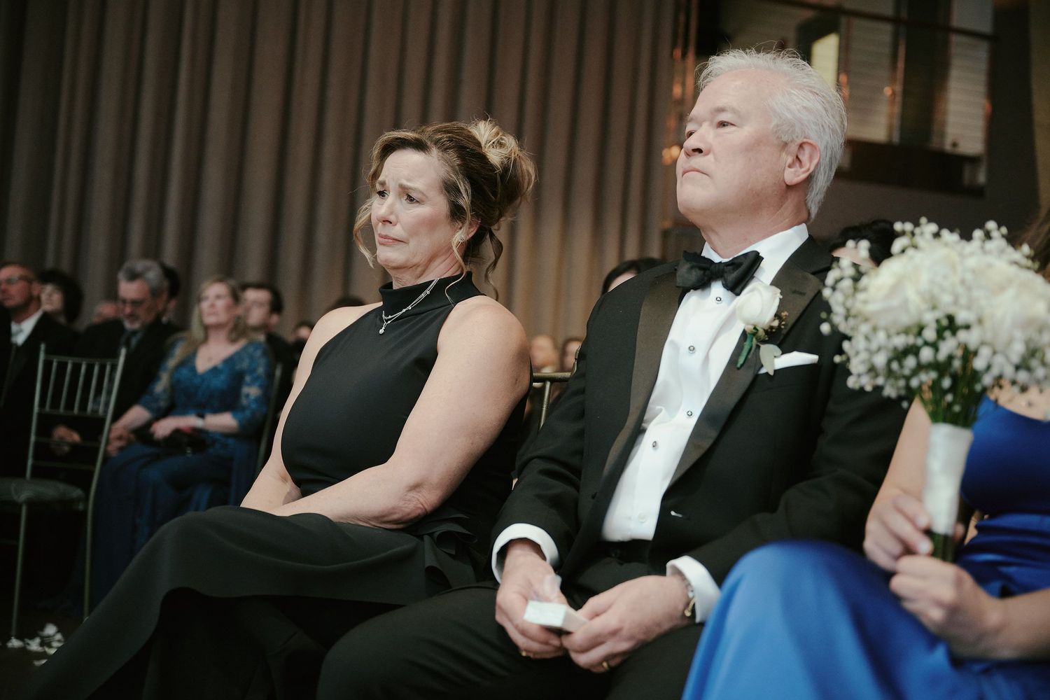 Two people in formal attire seated at an elegant event with floral decorations and black chiavari chairs.