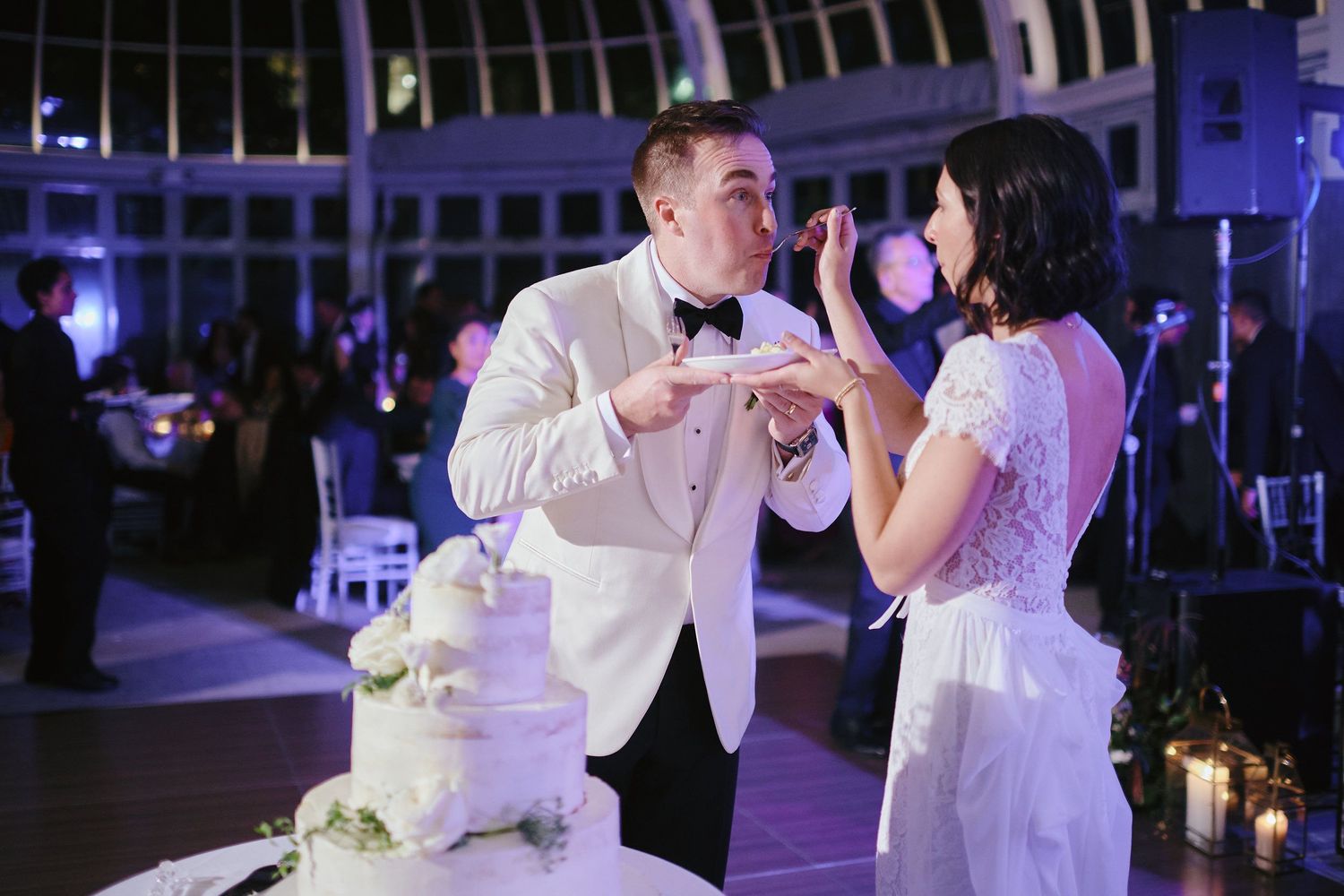 A couple in formal wedding attire laugh together while cutting their white wedding cake at an elegantly lit reception.
