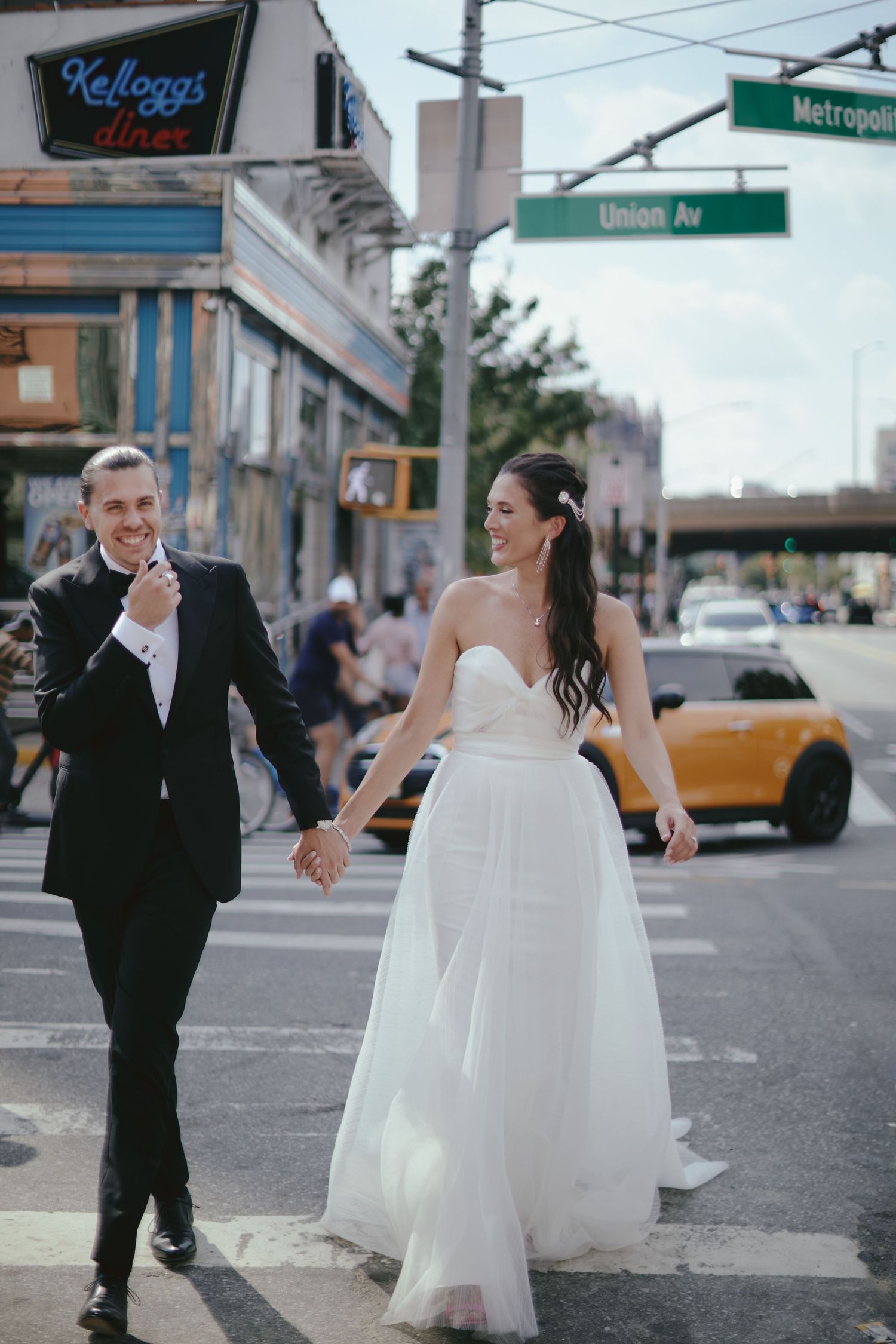 A couple in wedding attire holds hands while crossing a city street with distinctive signage and yellow taxi visible.