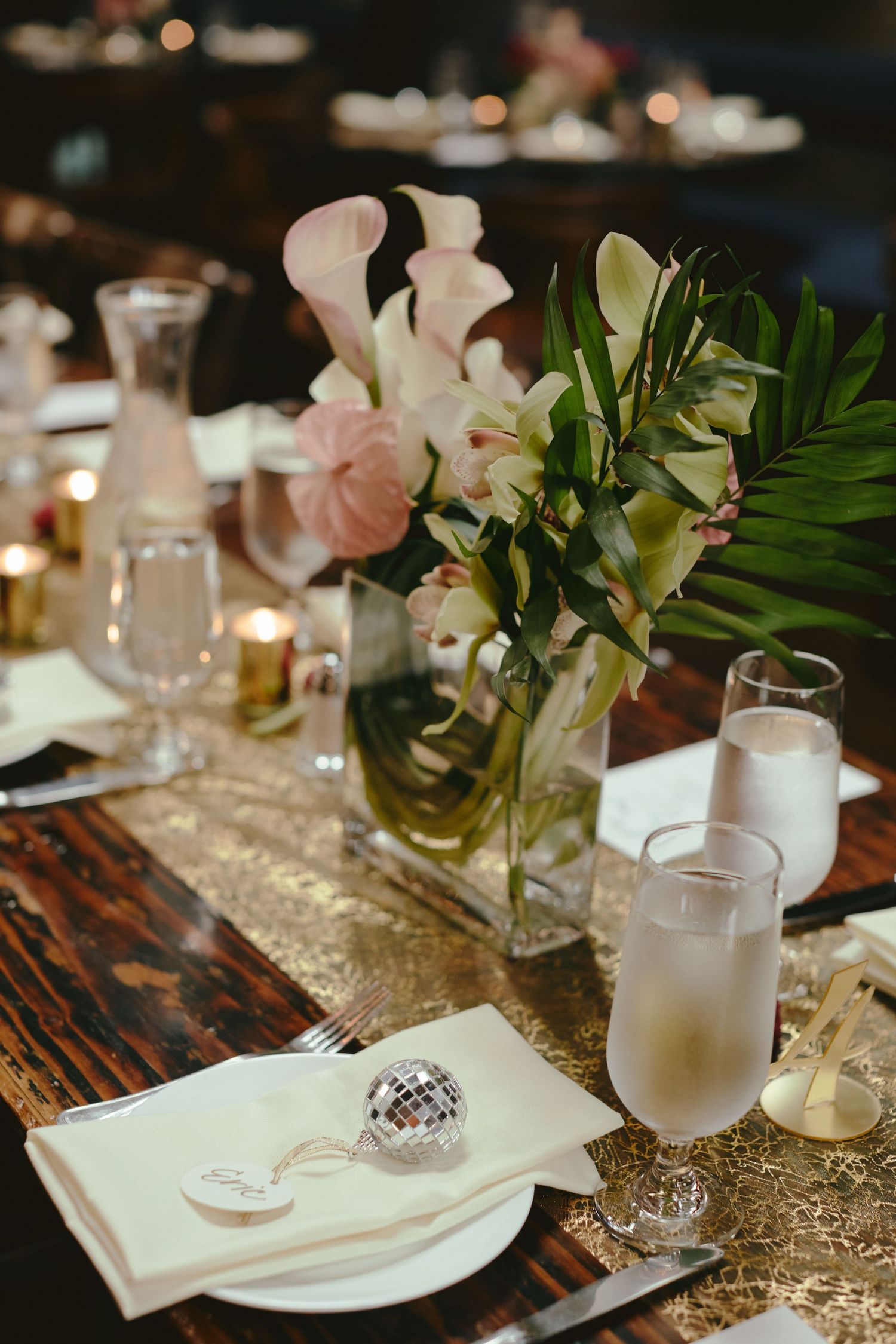 Elegant table setting with pink and white flowers in glass vase, frosted glasses and gold table runner on wooden surface.