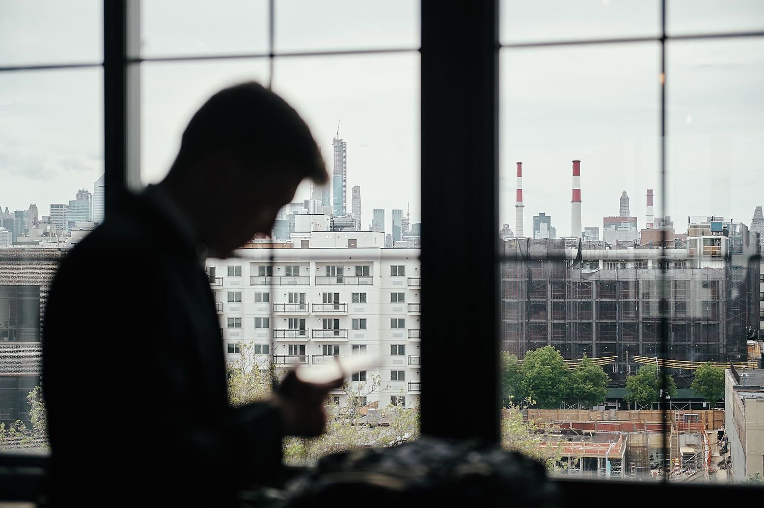 A silhouetted figure checking their phone by a window overlooking an urban cityscape with apartment buildings.