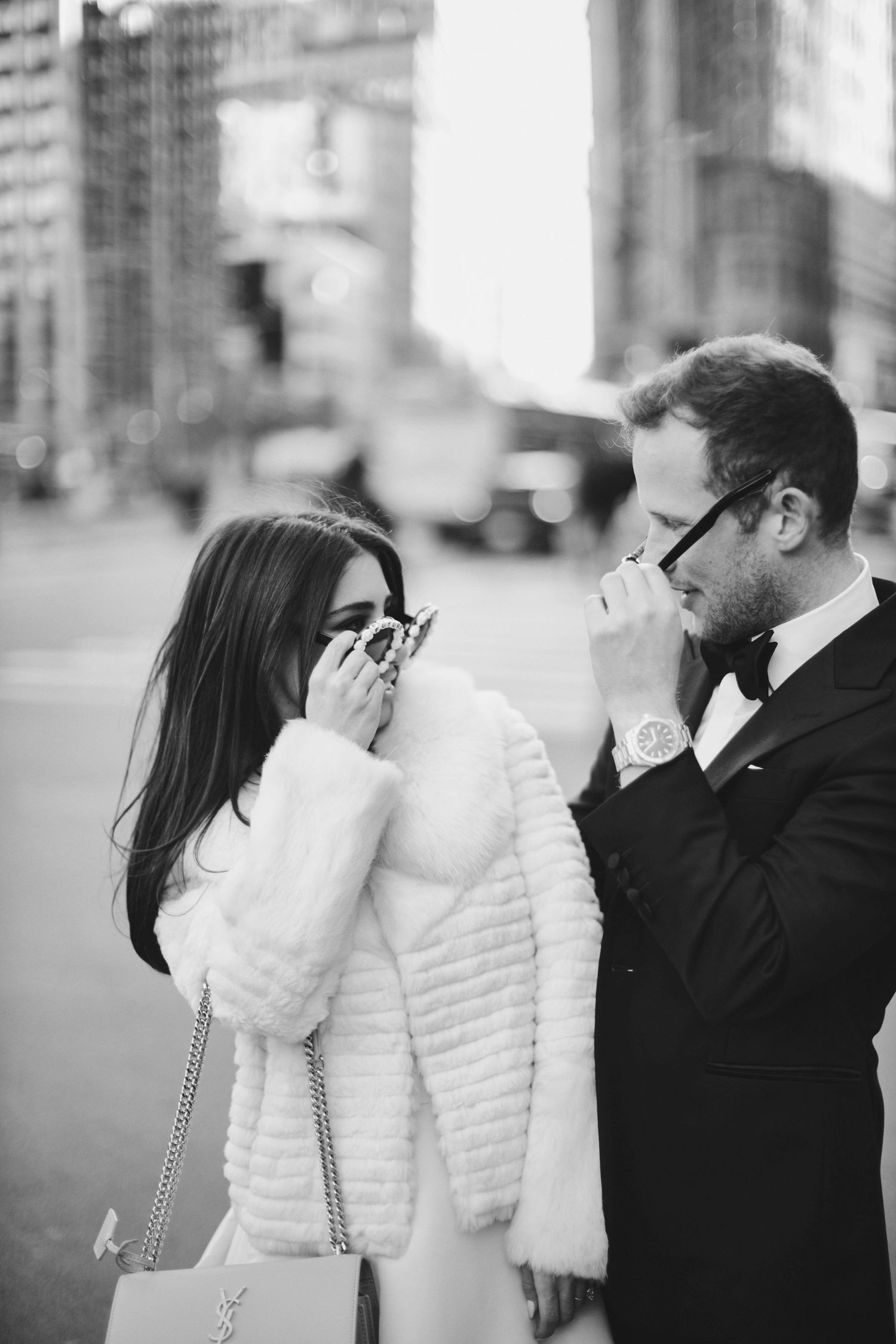 A couple in formal attire shares a candid moment on a city street, with her in a white fur coat and him in a bow tie.