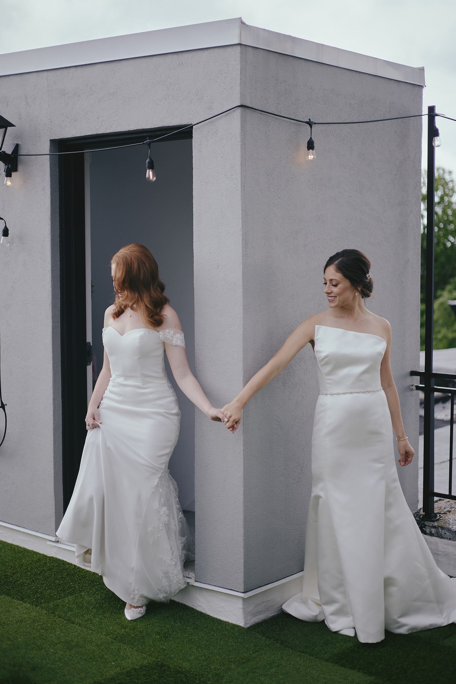 Two brides in elegant white wedding dresses hold hands while walking outside a grey building.