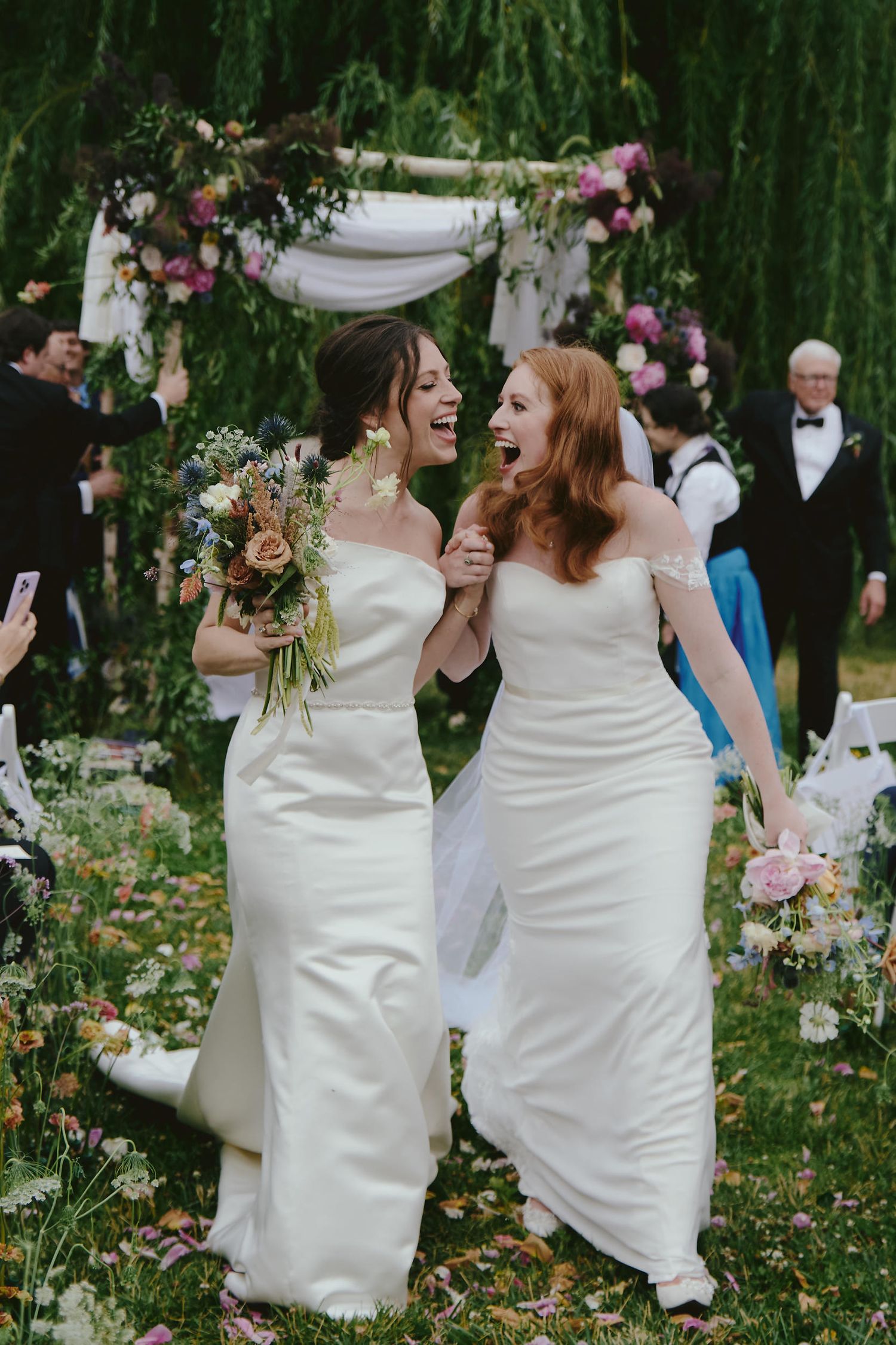 Two brides in white dresses share an ecstatic moment of laughter under a floral arch during their outdoor ceremony.