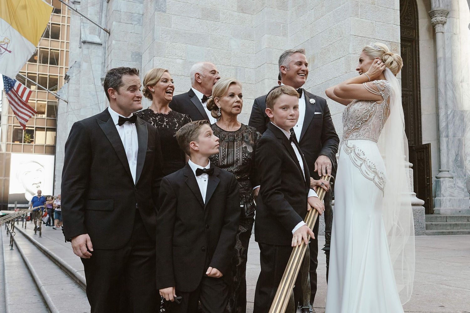 Wedding party members in formal attire gather together on stone steps outside an elegant building.