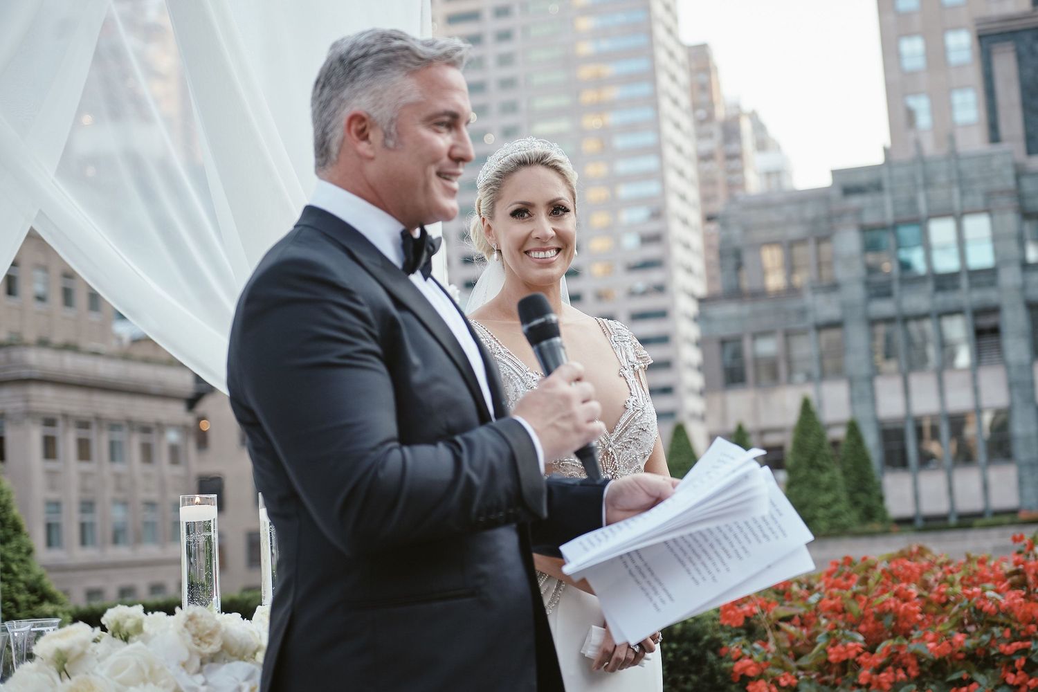A newlywed couple speaks during an outdoor ceremony in front of red flowers with urban buildings in the background.