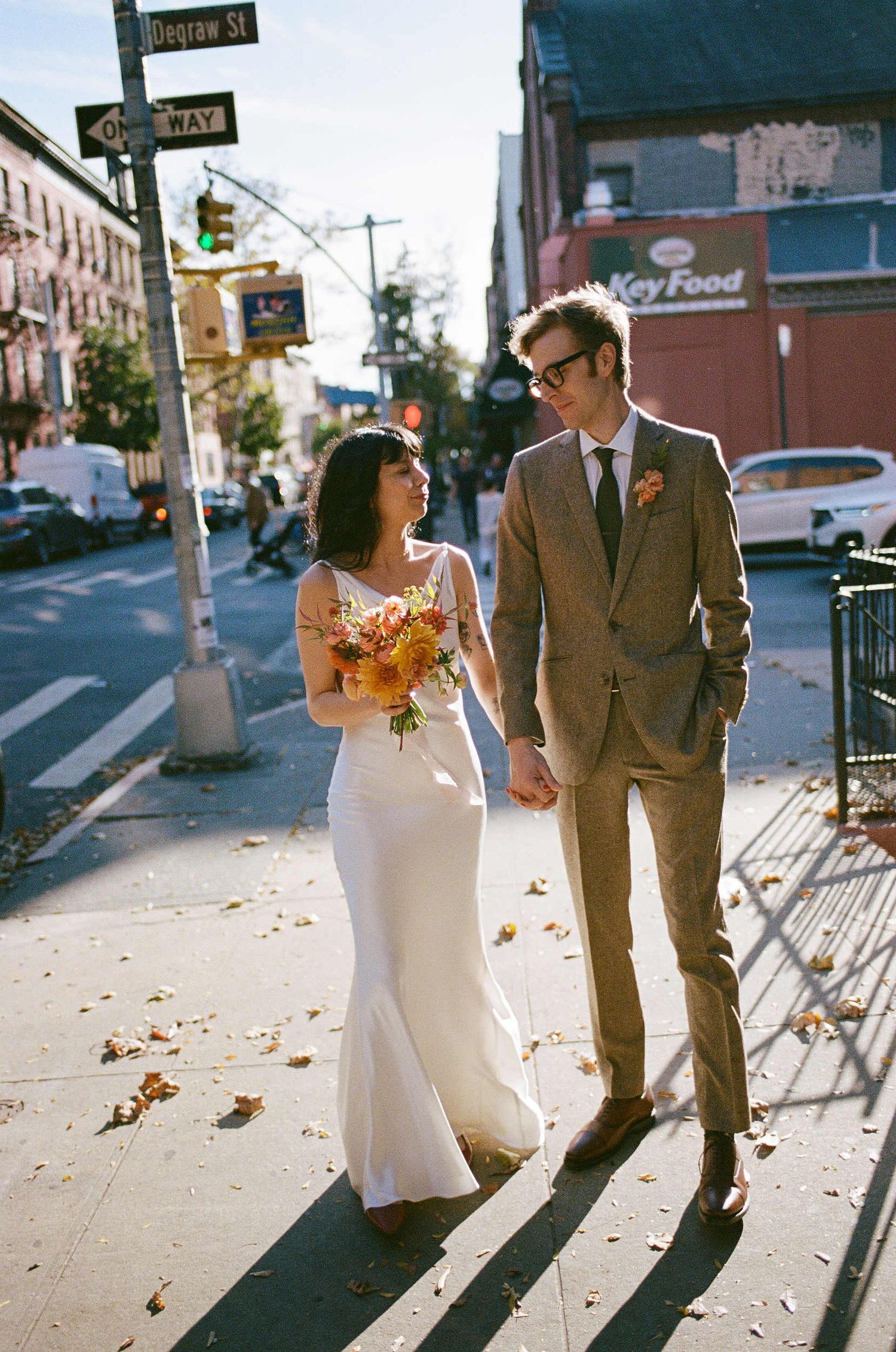 A stylish couple walks down a city street on their wedding day with the bride holding autumn-colored flowers.
