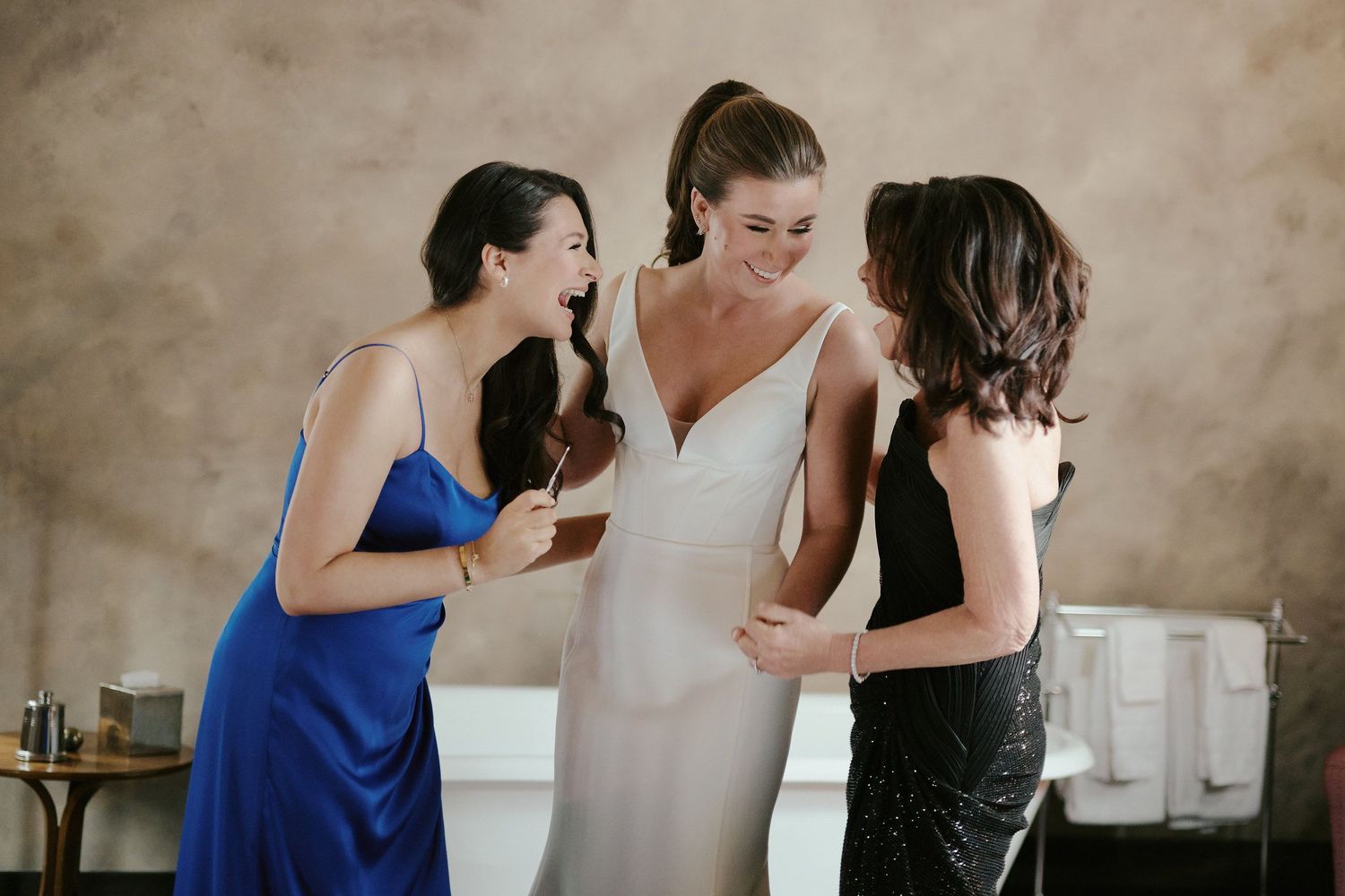 Three women share an intimate candid moment of laughter while getting ready for a formal event.