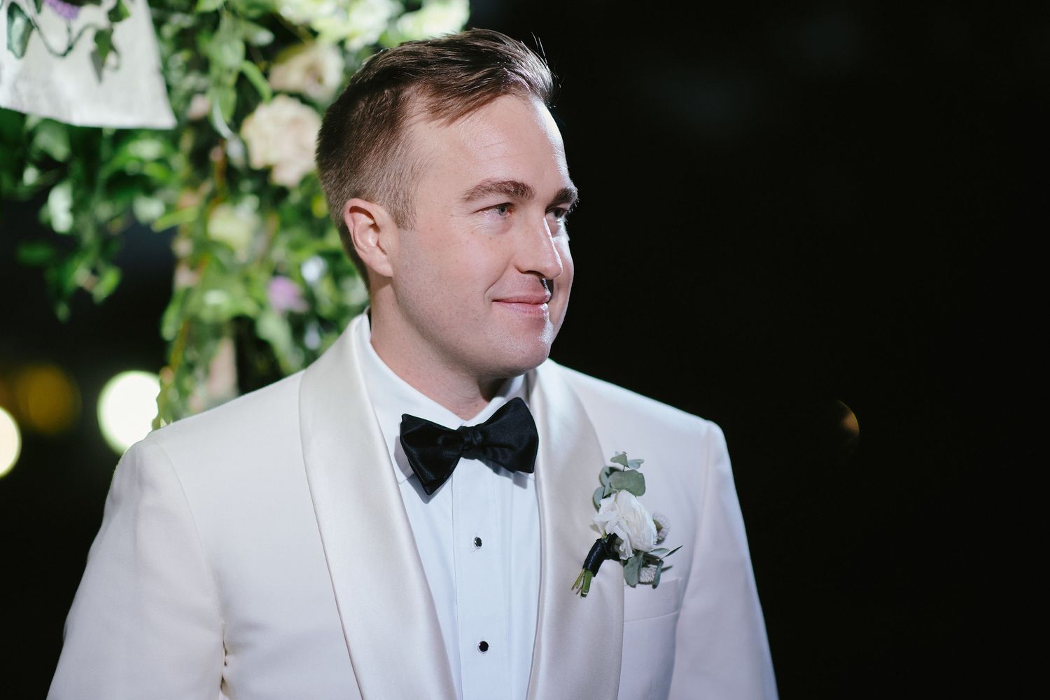 A groom in a white tuxedo jacket and black bow tie smiles while wearing a boutonniere at an evening wedding.