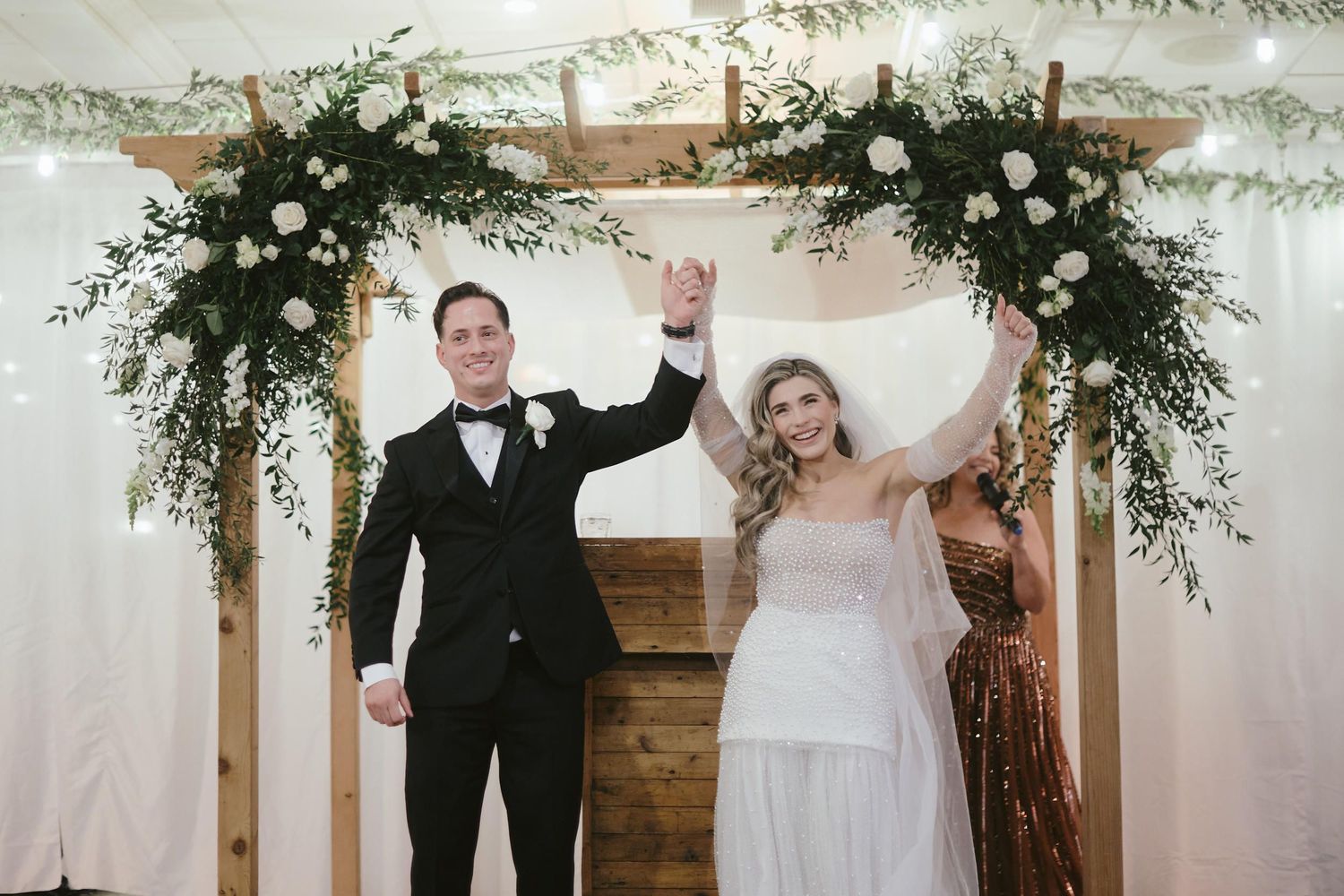 A joyful wedding celebration moment under a wooden arch decorated with lush white floral arrangements and greenery.