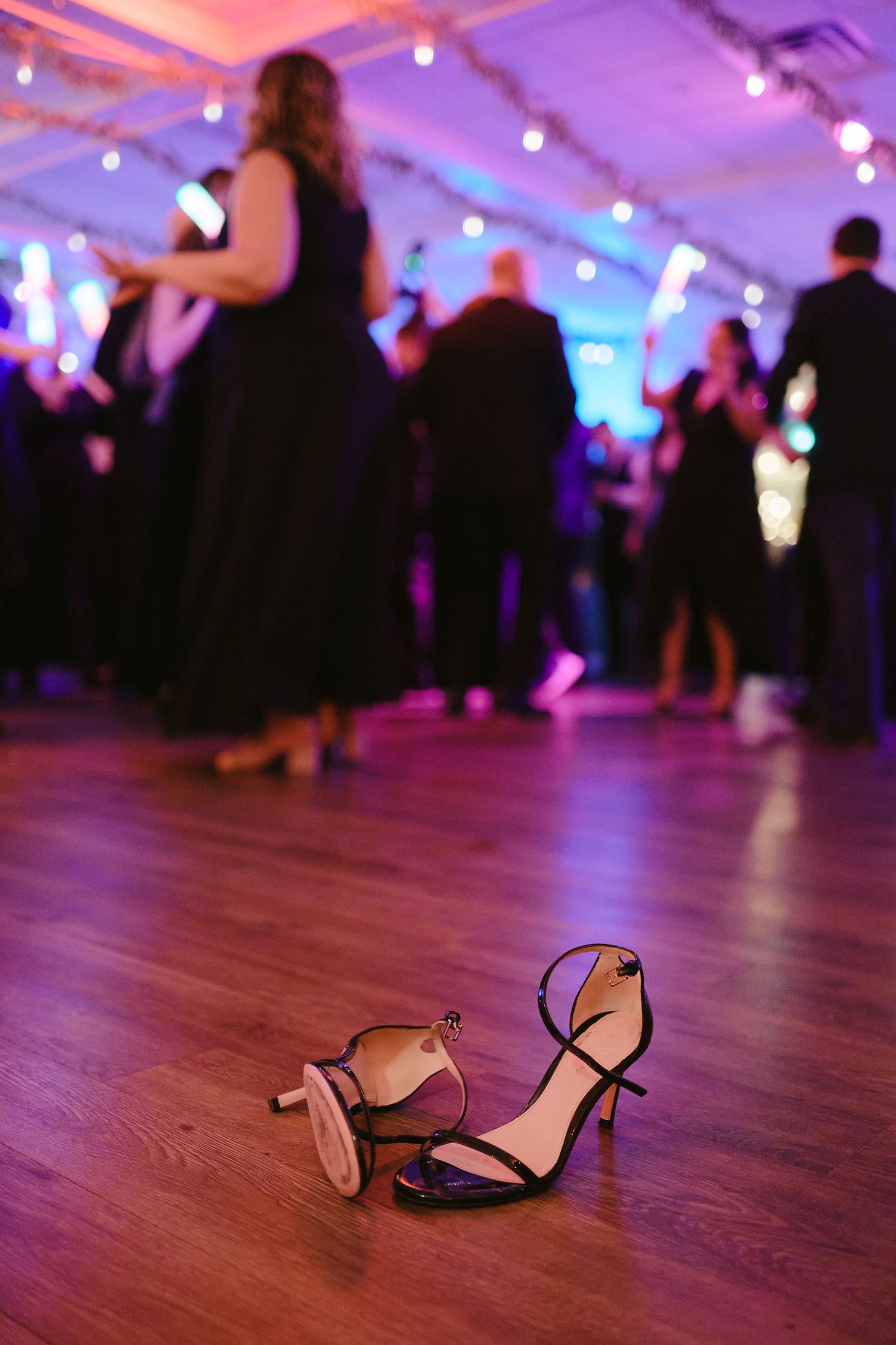 Abandoned high heels on a wooden dance floor with dancers silhouetted in purple lighting at a party.