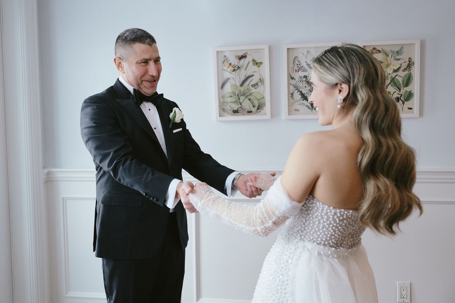 A couple shares a tender first look moment in an elegant white room with framed botanical prints on the wall.