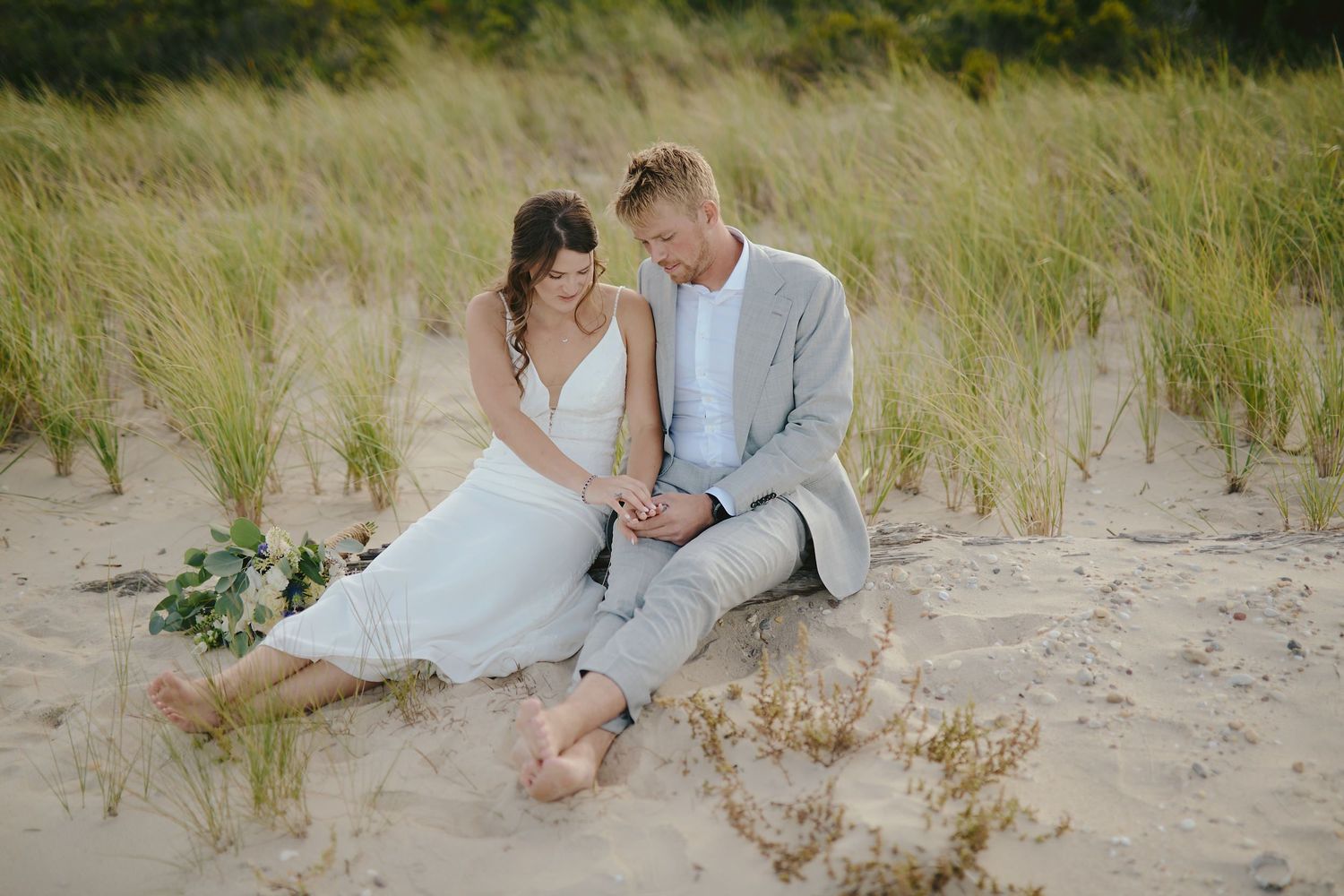 A romantic couple in wedding attire sit together on sandy dunes with tall grass in the background.