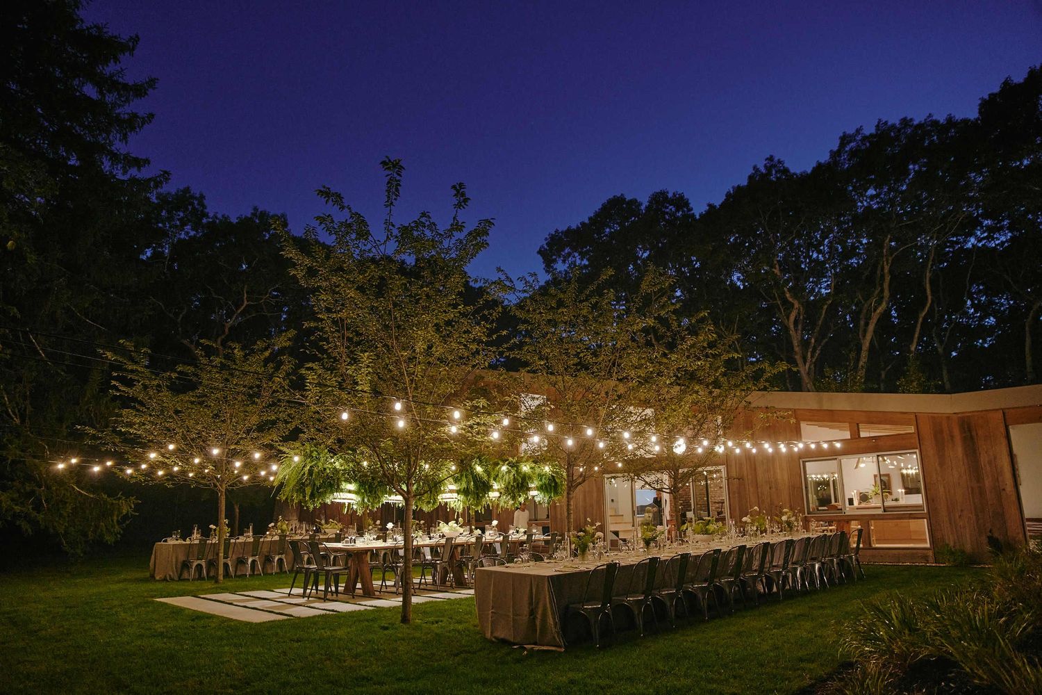 Outdoor wedding reception setup with string lights illuminating long tables under a dark purple evening sky.