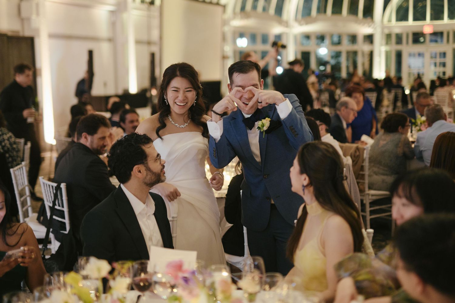 Bride and groom laughing together at elegant wedding reception dinner with guests seated at floral-decorated tables.