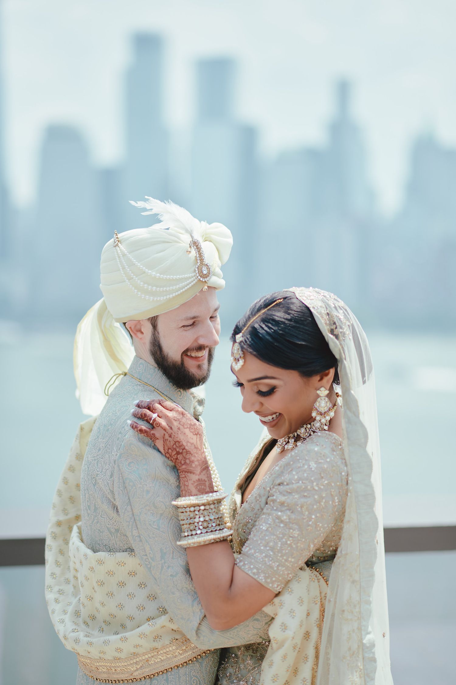 A romantic wedding couple in traditional Indian attire embraces against a city skyline backdrop.