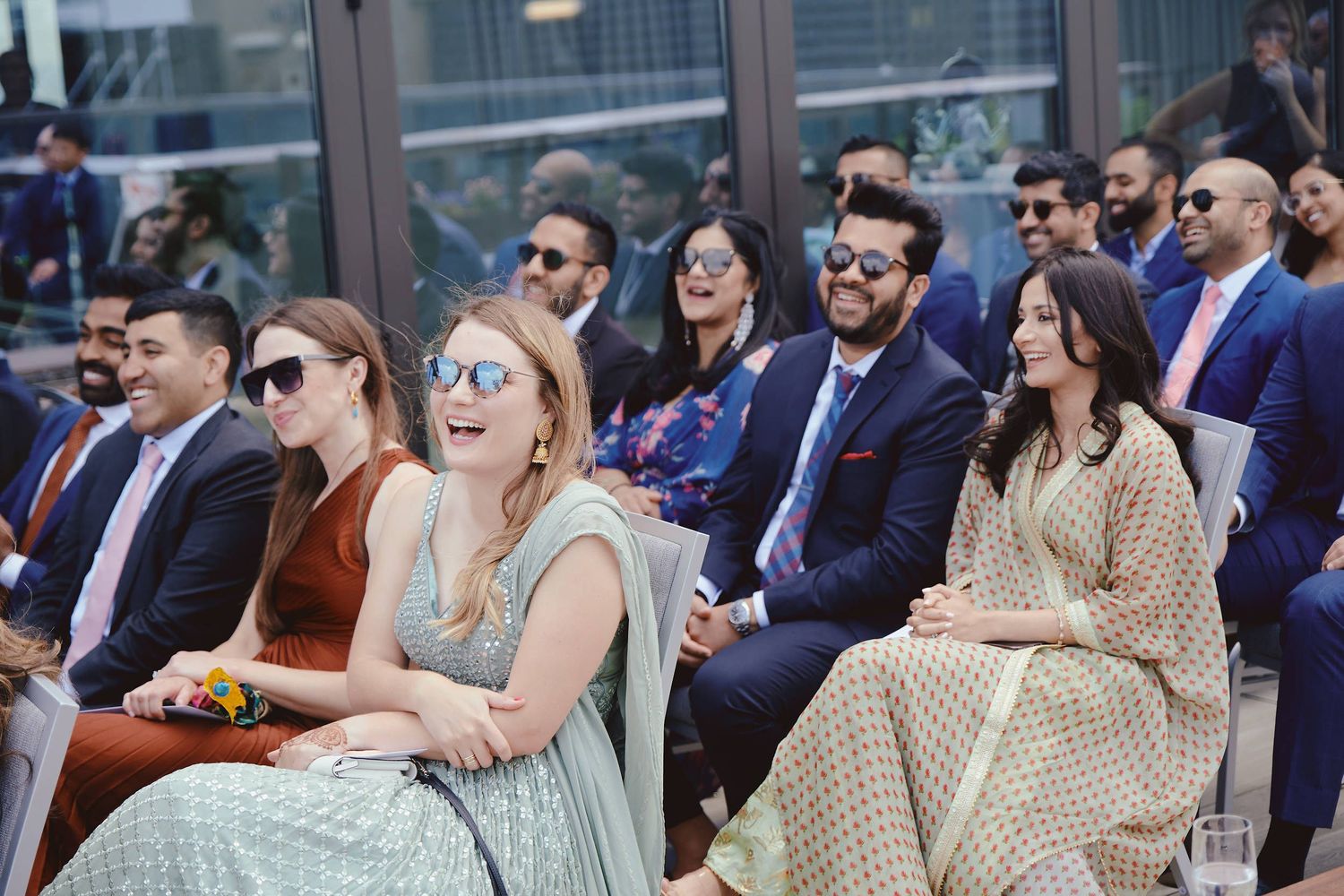 Wedding guests seated outdoors at a ceremony wearing formal attire and sunglasses on a sunny day.
