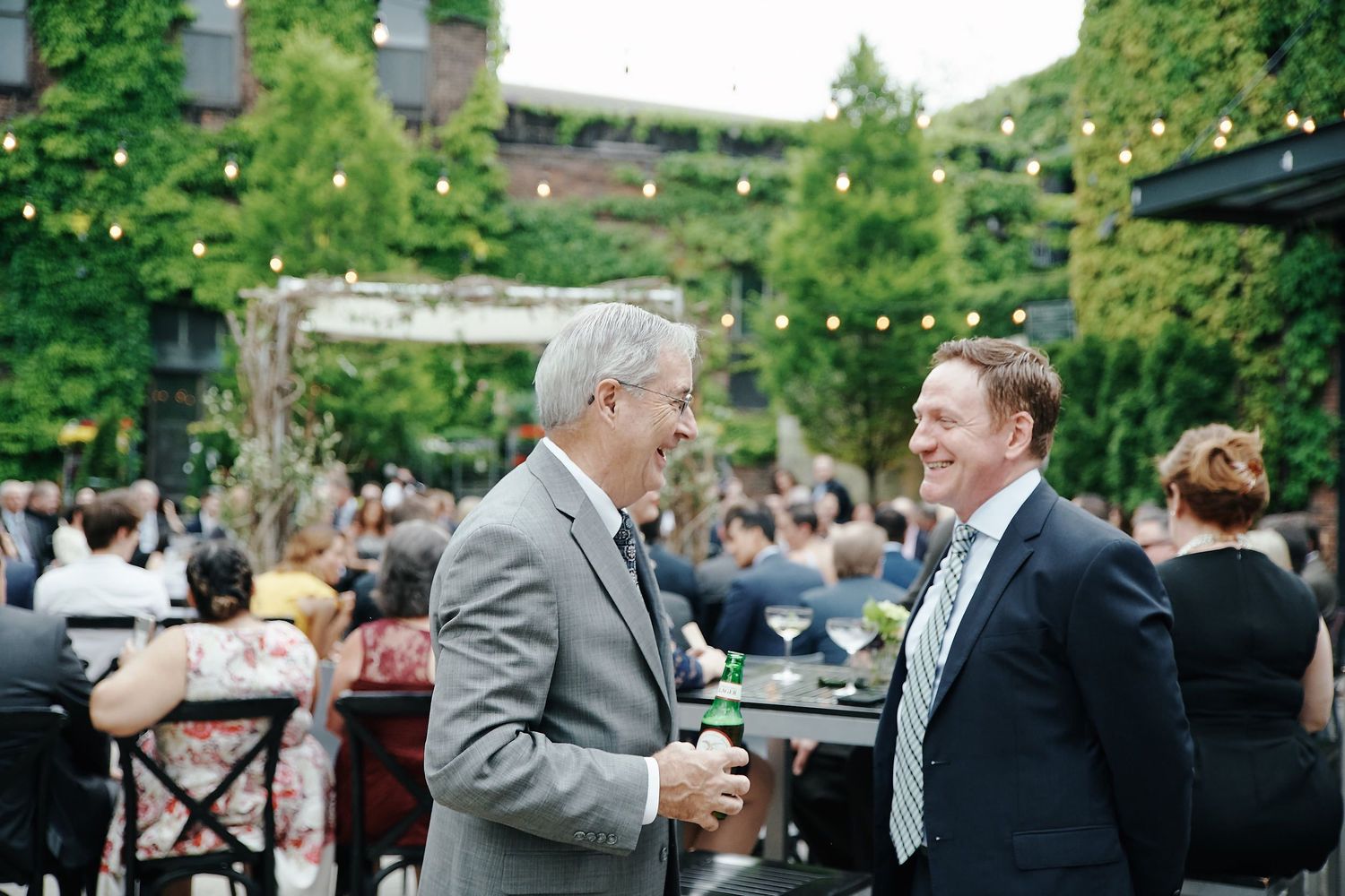 Two men in suits having an animated conversation at an outdoor garden party with guests in the background.