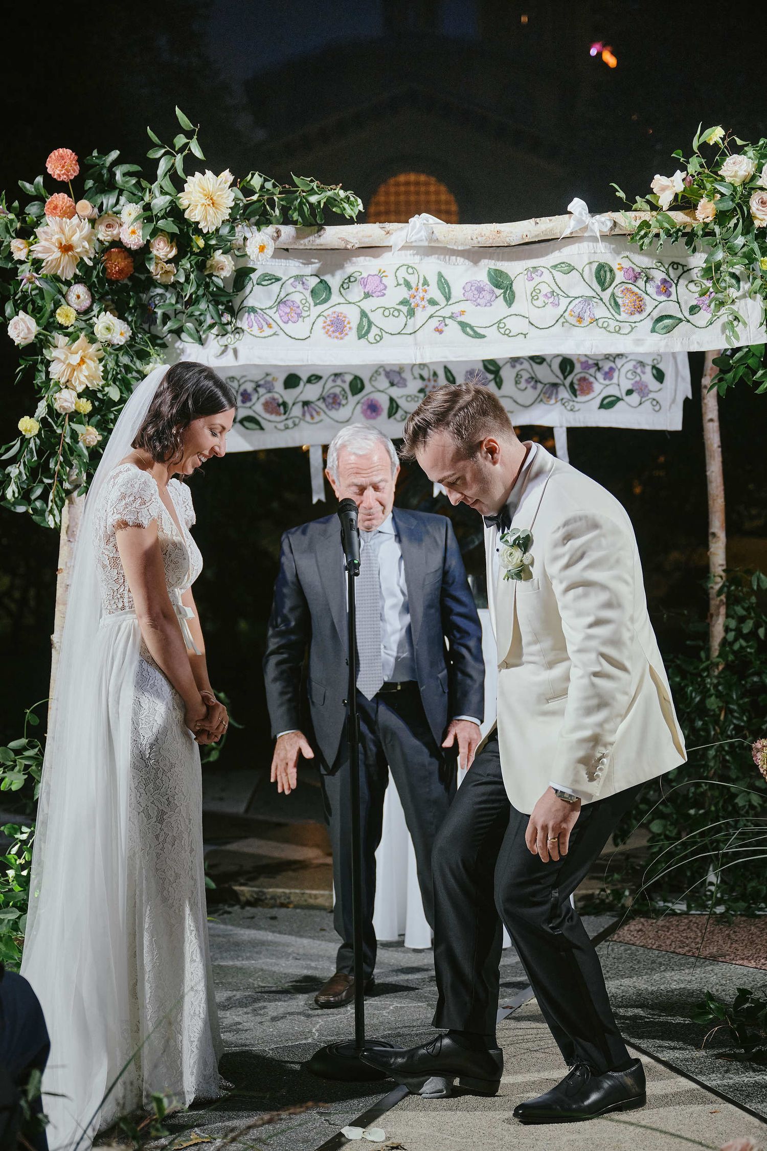 A wedding ceremony takes place under a floral arch with white lace and greenery decorations at night.