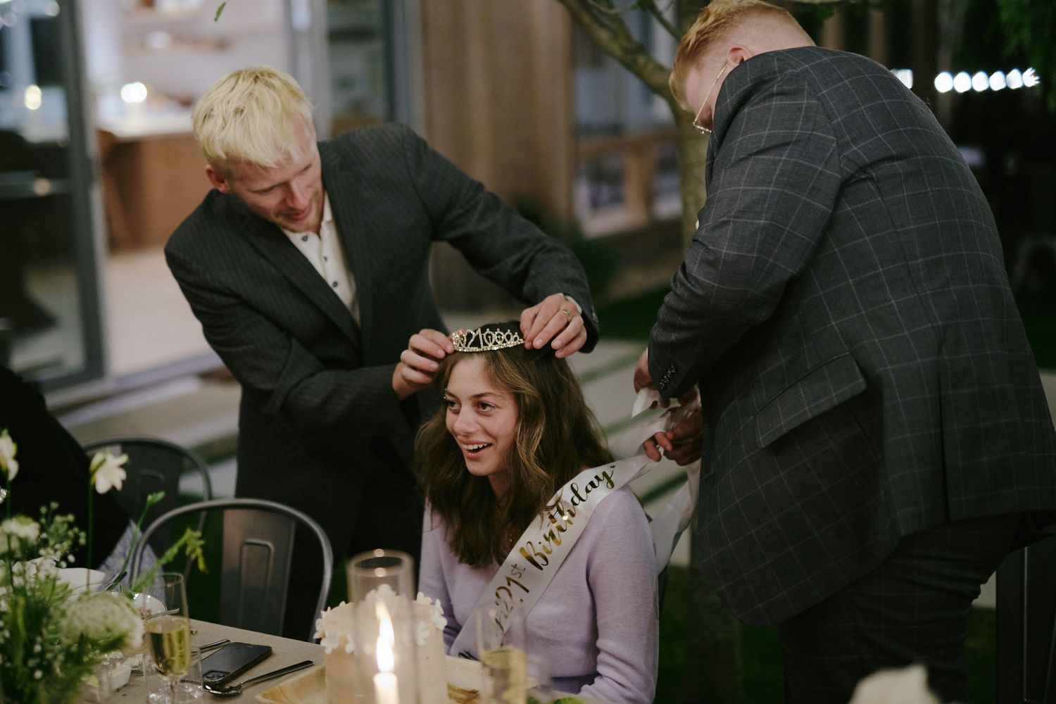 People in dark outfits assist someone in a lavender sweater at an intimate candlelit table setting with greenery.