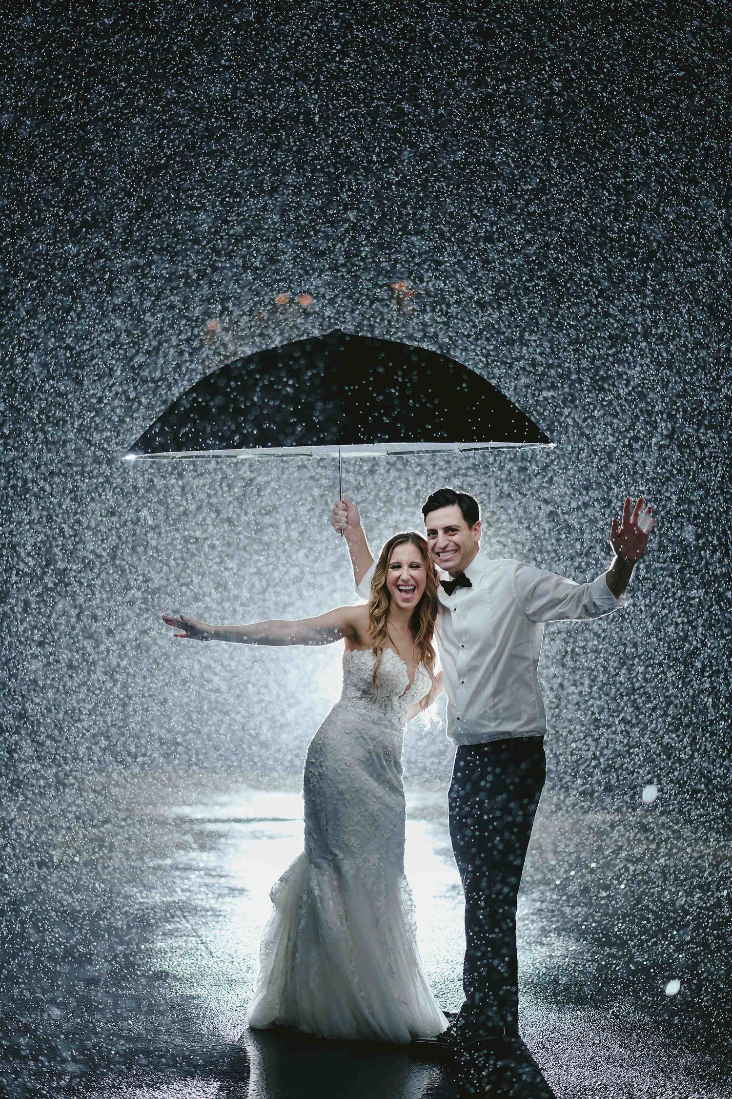 Couple embraces under an umbrella in the rain during a romantic wedding photoshoot.