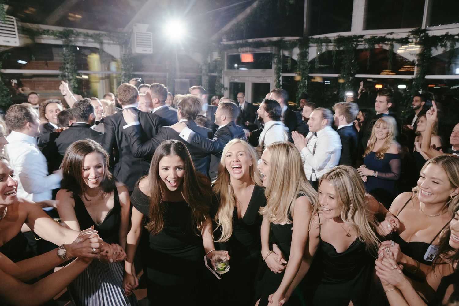 Crowded nightclub scene with partygoers in elegant black dresses and formal attire dancing and socializing under bright lights.