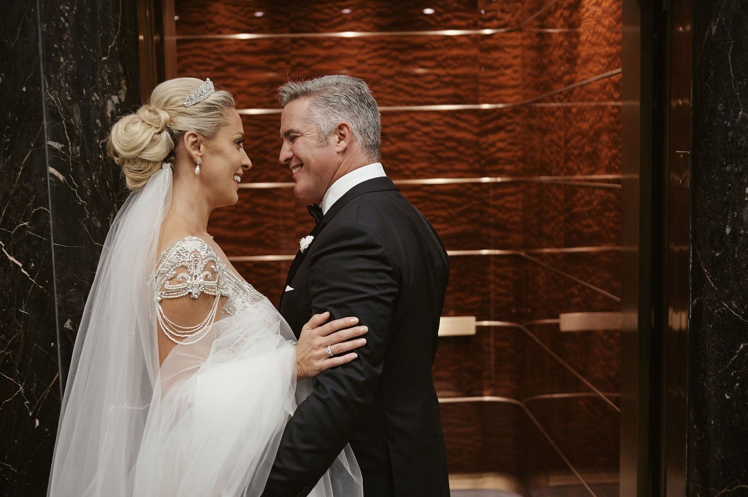 A bride and groom share an intimate moment in a warmly lit hallway with dark wood paneling.